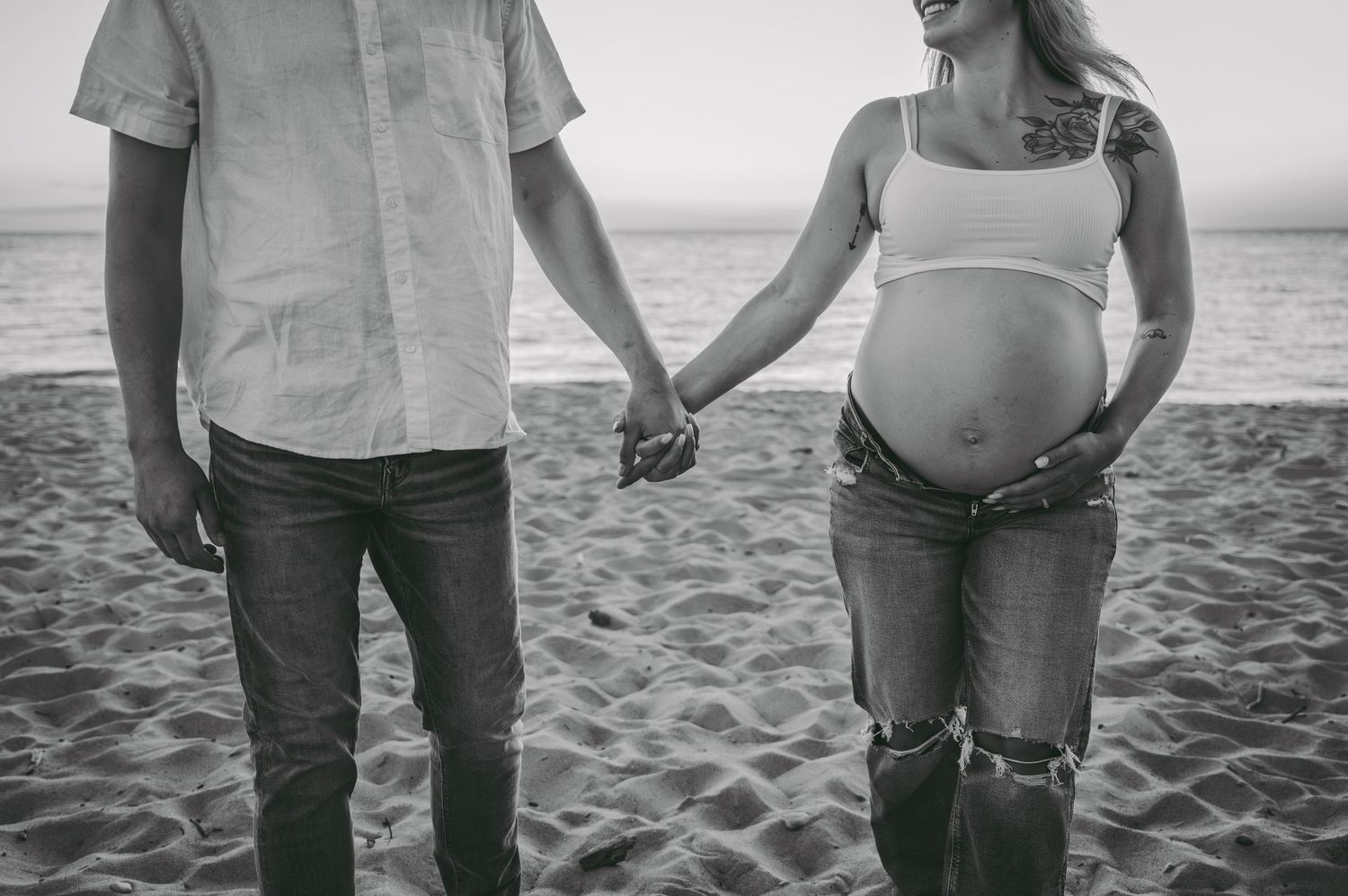 Black and white photo of a couple holding hands on a beach, showing a pregnant belly in casual attire.