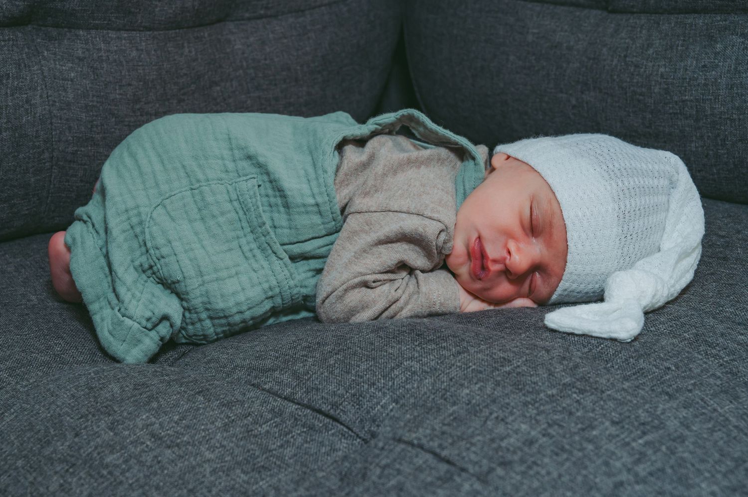 Peaceful sleeping newborn in green and gray clothing resting on a dark gray couch.