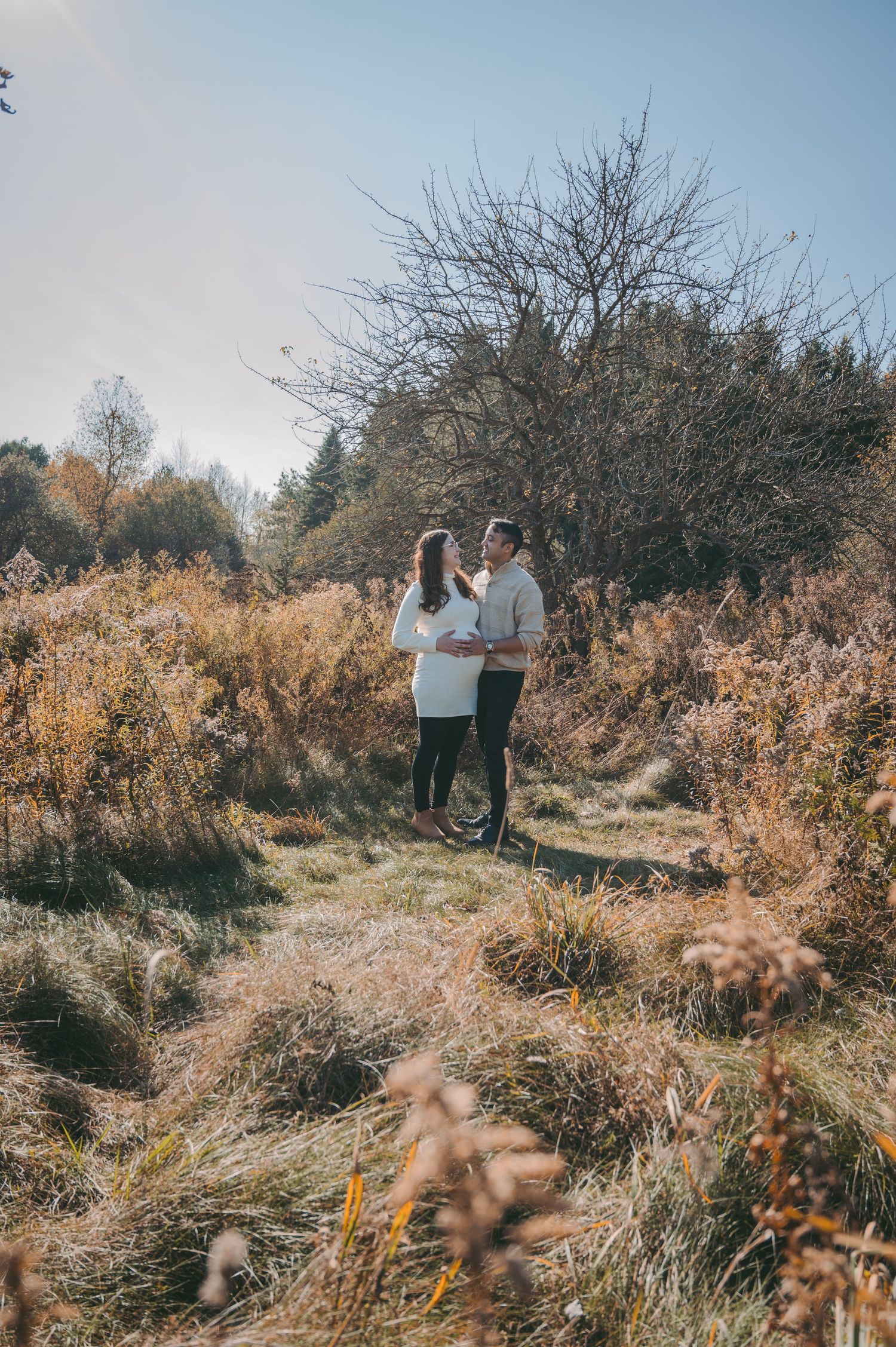 Couple standing together in a field of tall autumn grass and bare trees.