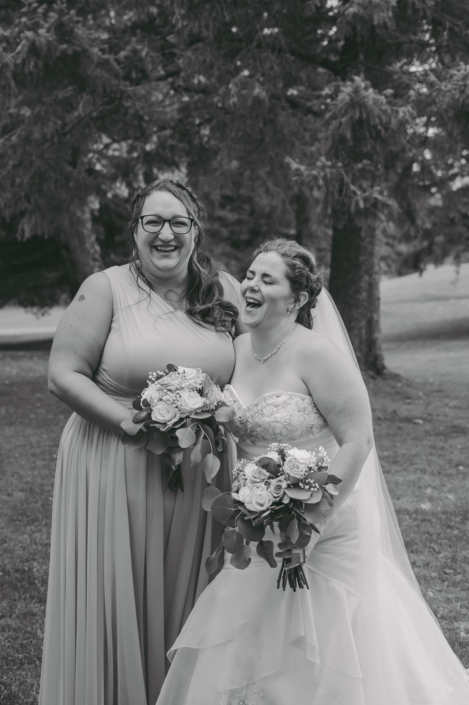 Bride and bridesmaid holding bouquets share genuine laughter in outdoor setting.