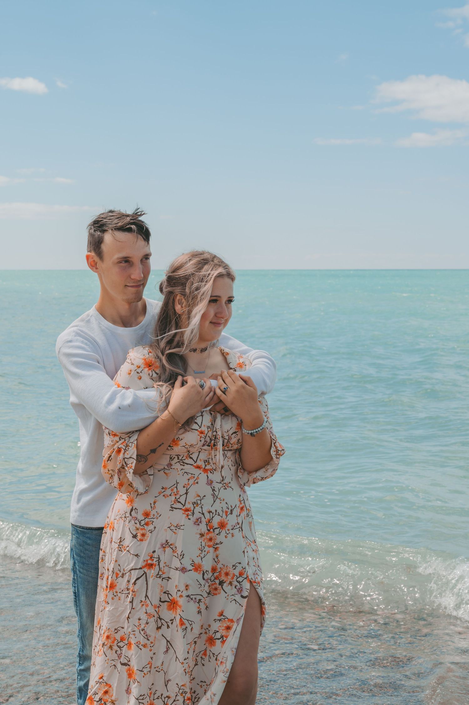 Tender moment between couple in floral dress and white sweater against turquoise ocean waters.