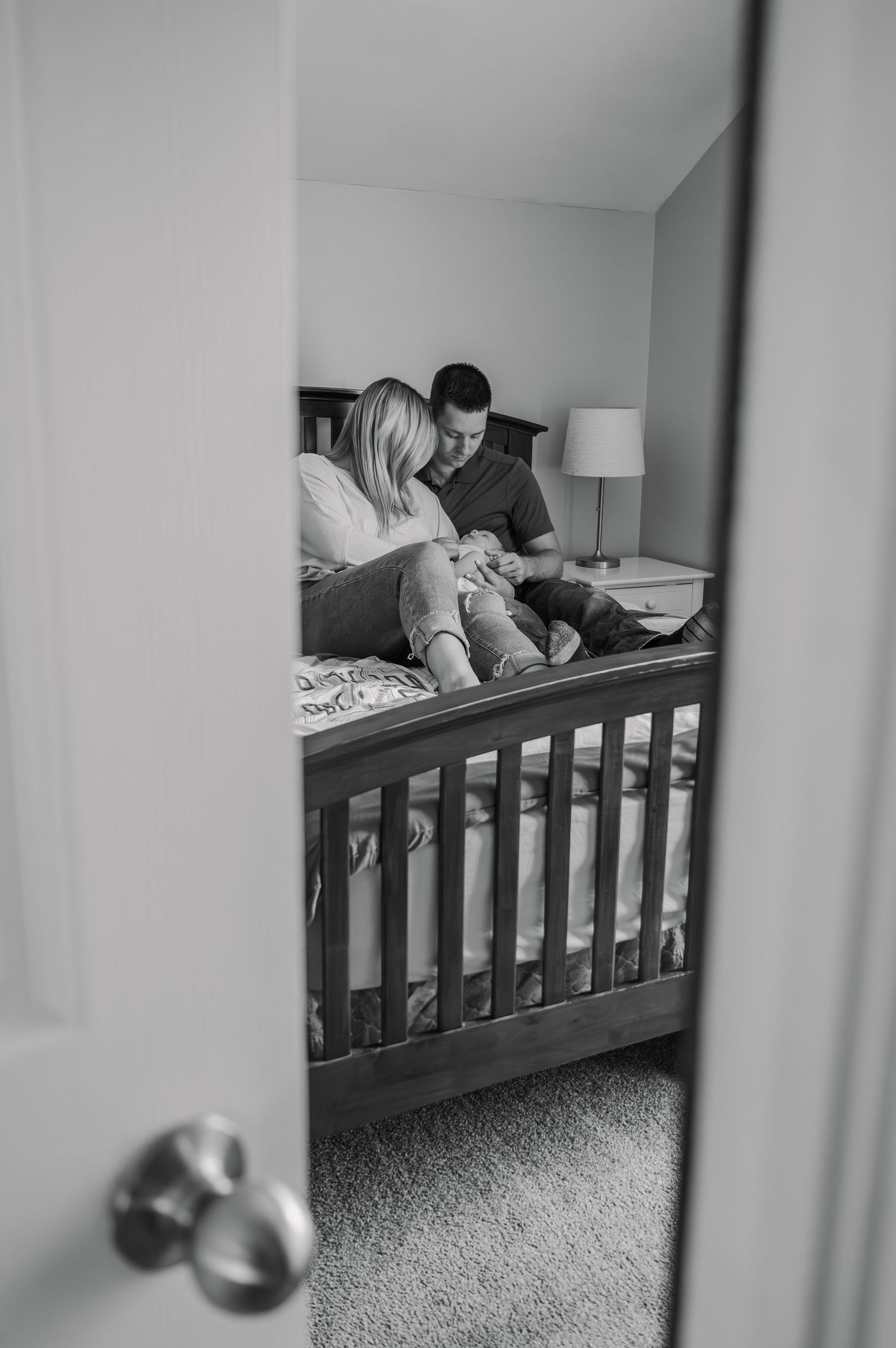 A black and white view through a doorway of a bedroom showing new parents snuggling their newborn on a bed with wooden railing.
