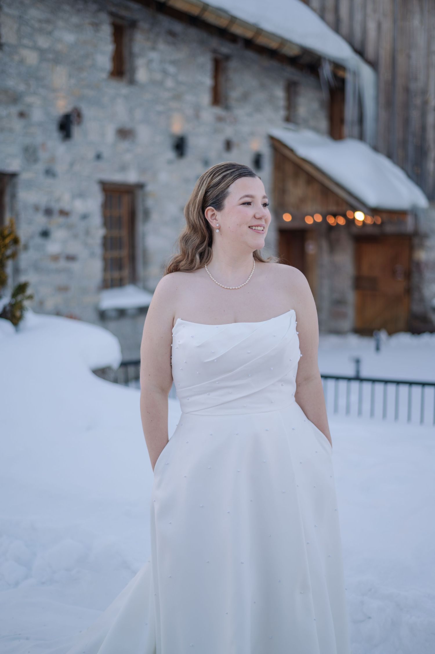A bride in a white wedding dress stands in front of a rustic stone building (Brussels Four Winds Barn) with snow on the ground.