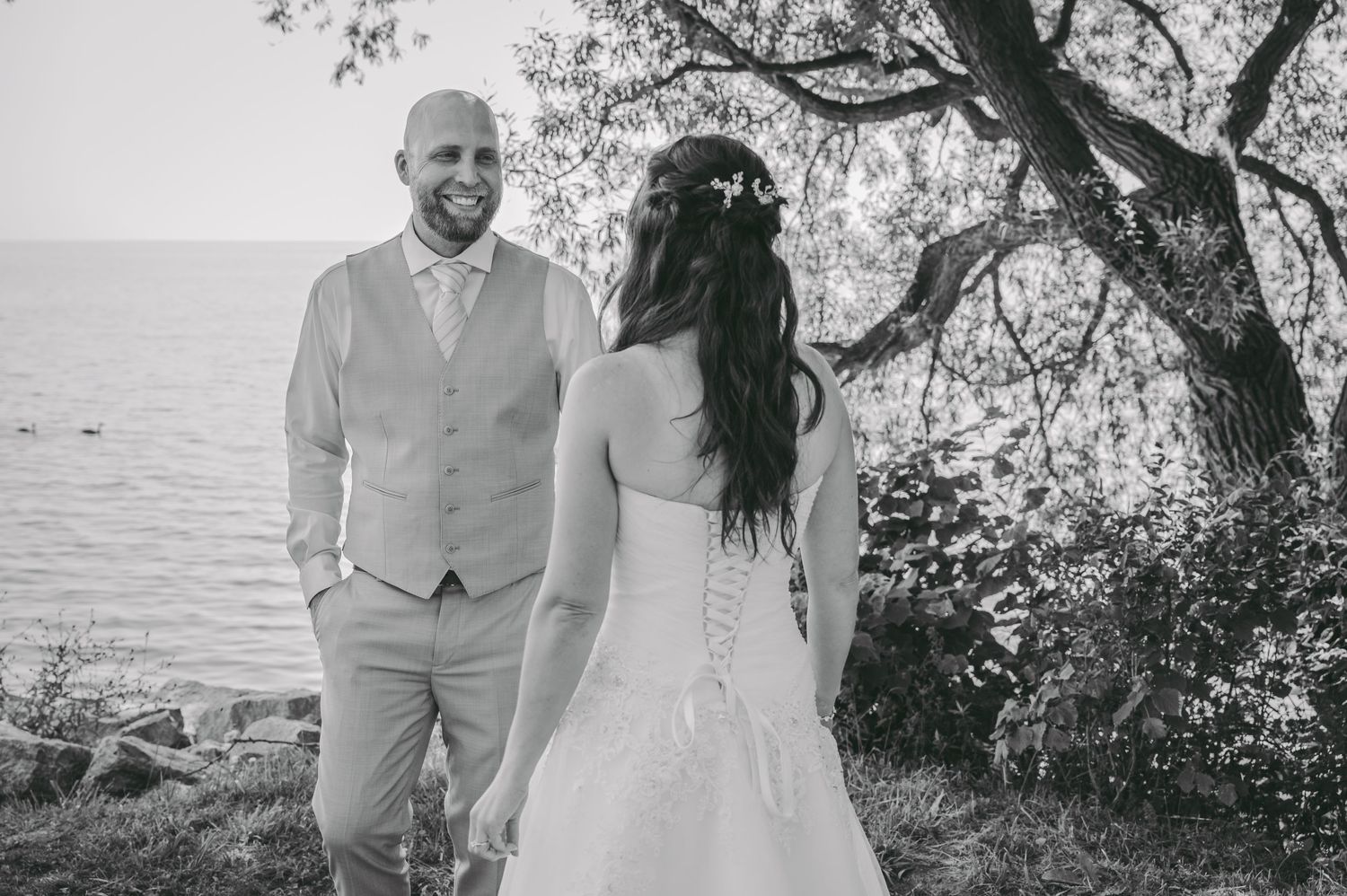 A couple in white wedding attire walks along a scenic beach path under trees at sunset.