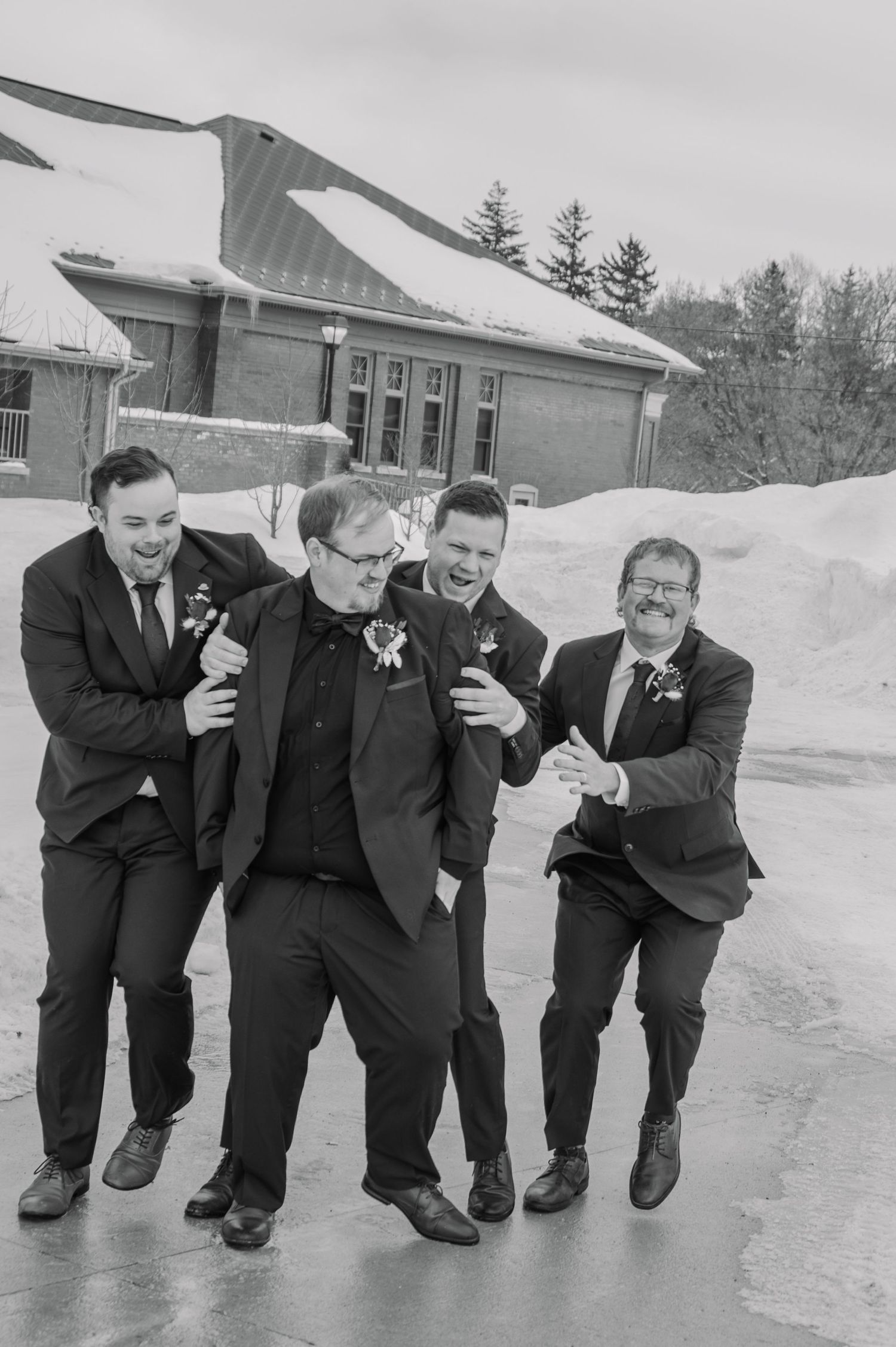 Black and white photo of groom and groomsmen laughing together outside a snowy winter wedding venue.