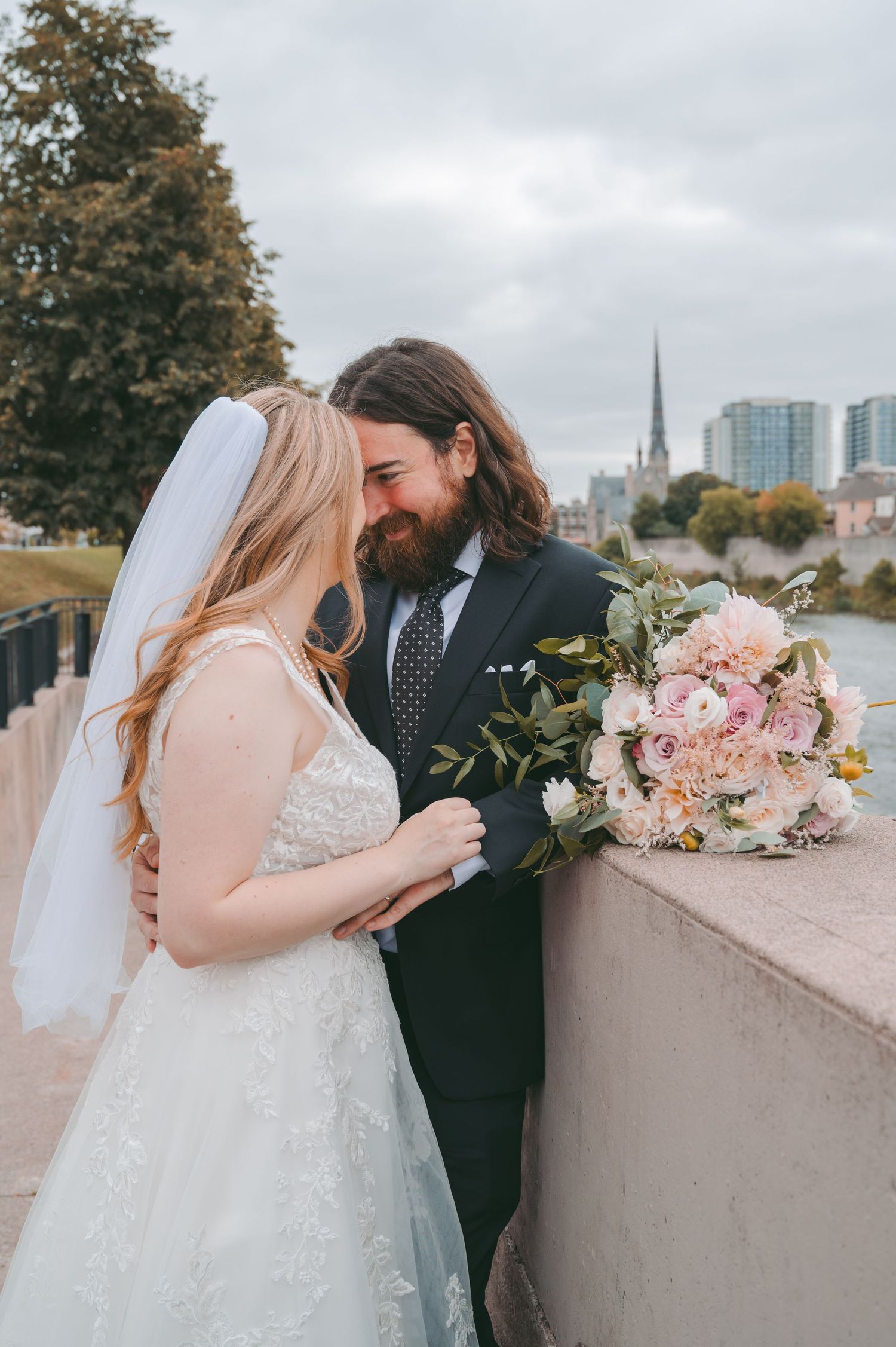 A romantic wedding couple shares an intimate moment by a stone wall with a beautiful floral bouquet and city skyline behind them.