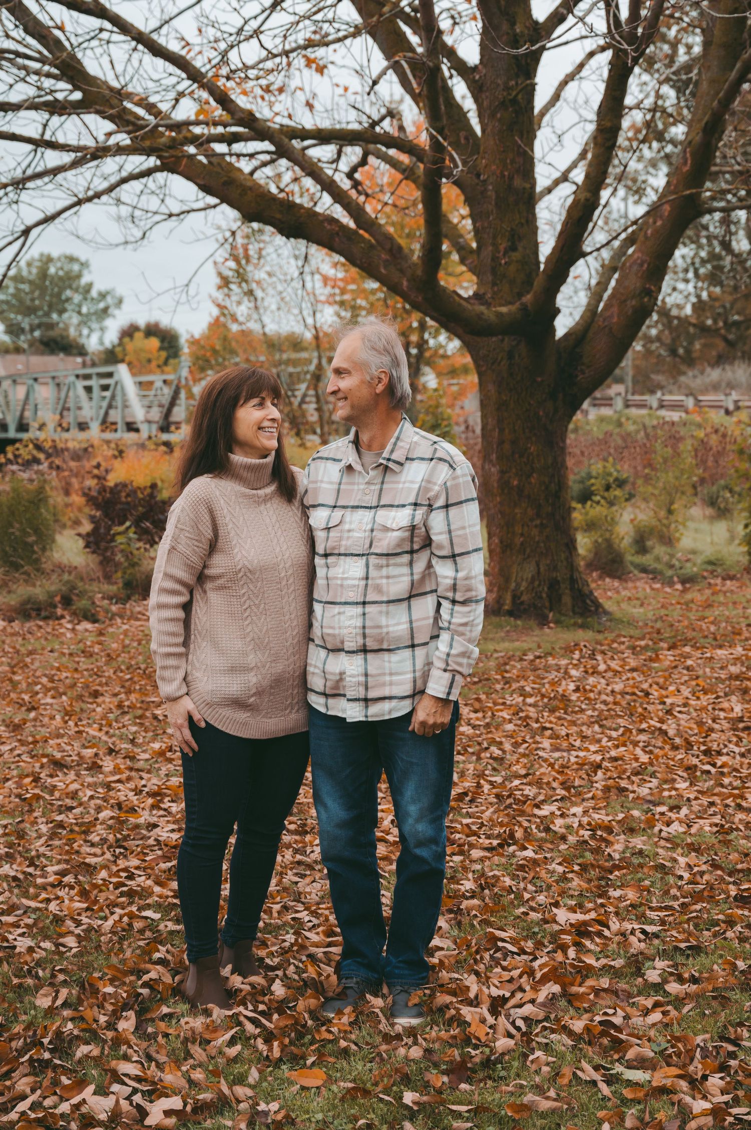 A couple walking through fallen autumn leaves under large trees in a park.