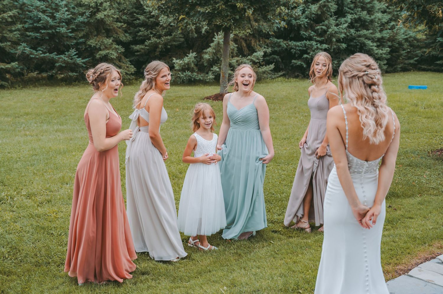 Wedding party in pastel colored dresses gathered on a green lawn with evergreen trees in background.