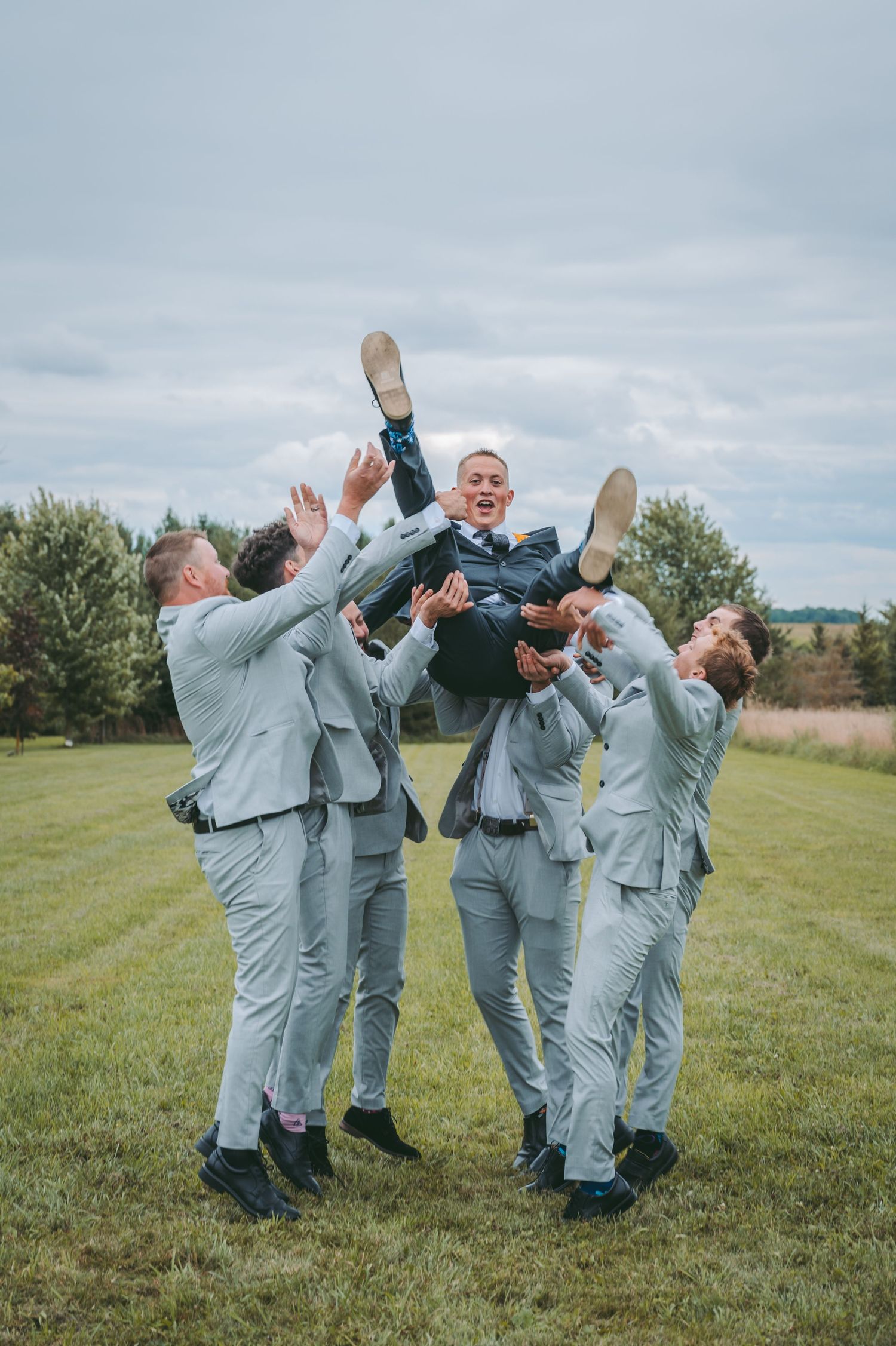Groomsmen in light gray suits tossing someone in the air during outdoor wedding photoshoot.