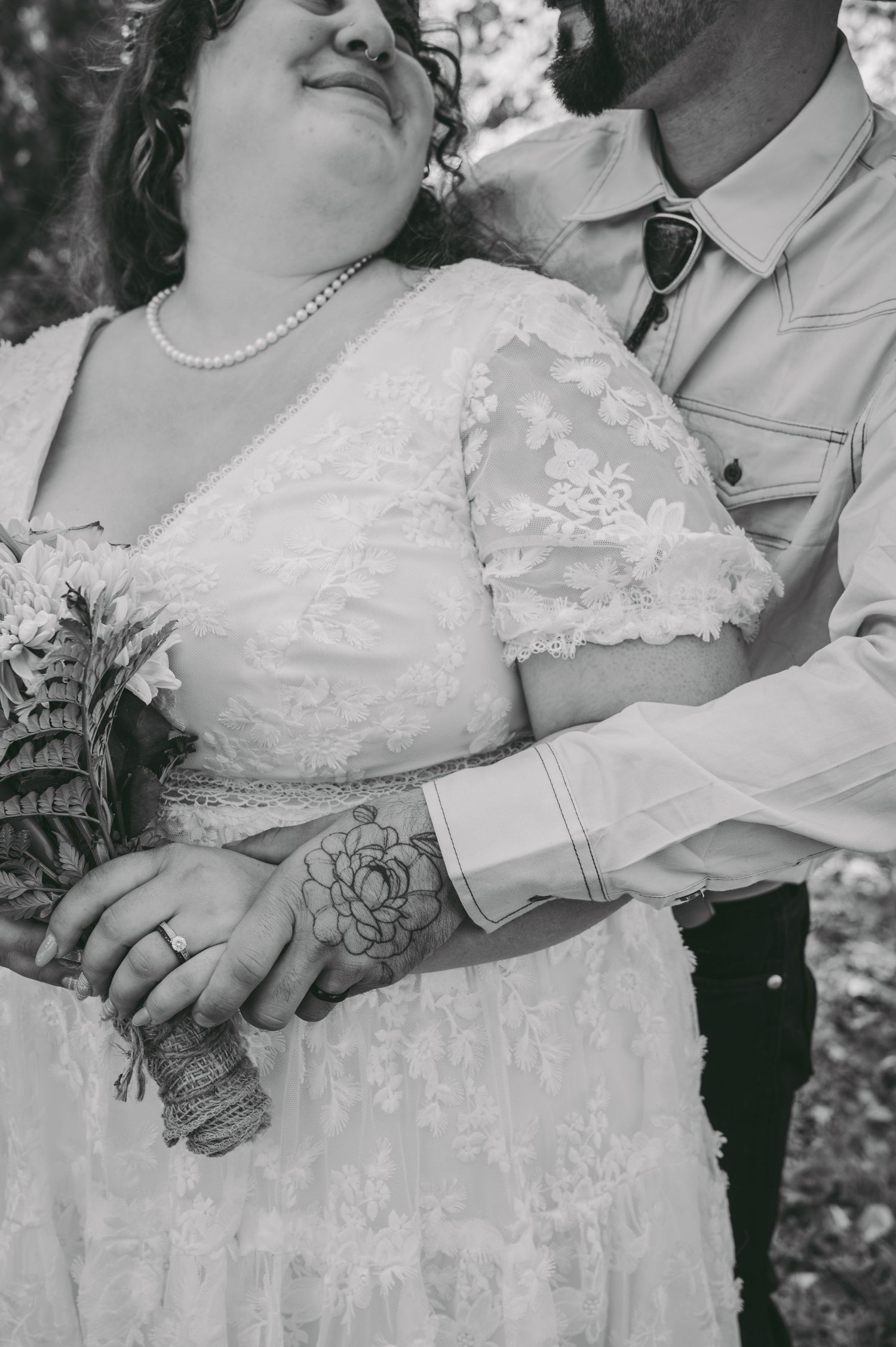 Close-up black and white detail of wedding dress lace and tattooed arms holding flowers.