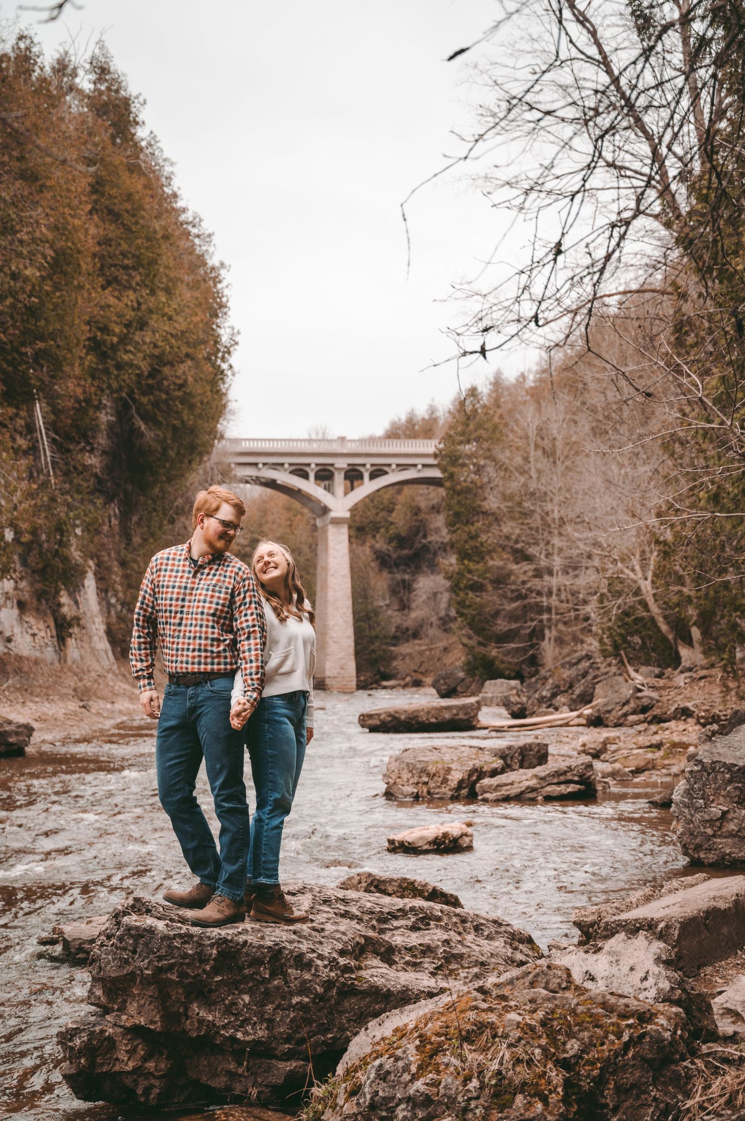 Couple standing on rocky riverbed with autumn foliage and bridge in background.