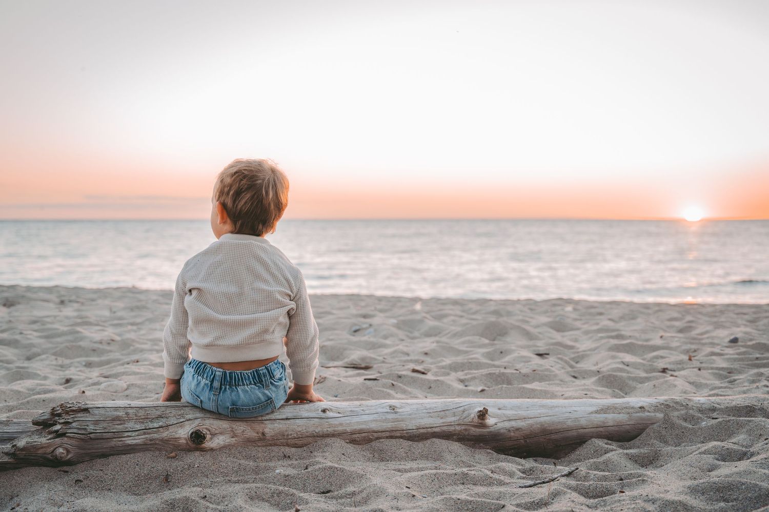 Child sits alone on sandy beach at sunset watching the horizon.