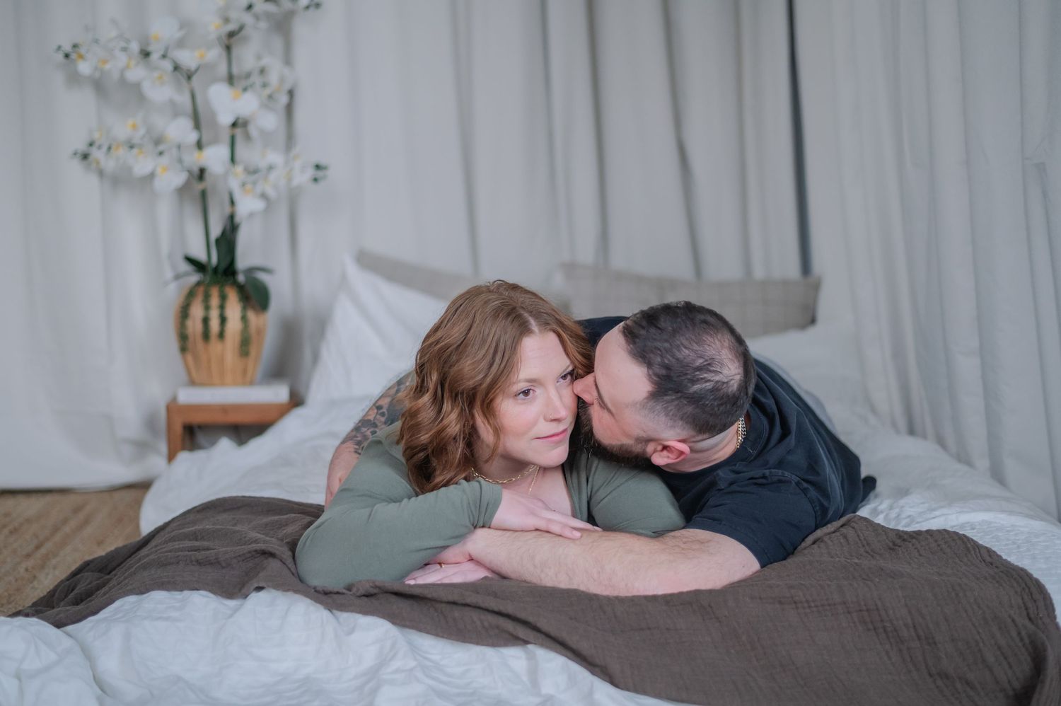 Couple lying on a bed, man kissing woman's cheek as she smiles, with white orchids in the background.