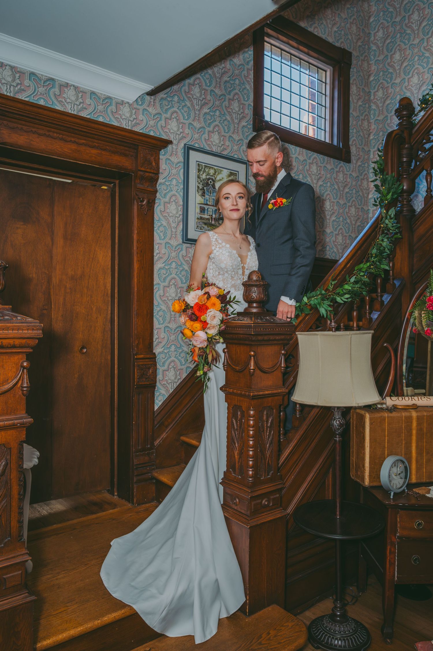 Bride and groom pose elegantly on a wooden staircase in a vintage mansion, bride holding a vibrant orange bouquet.