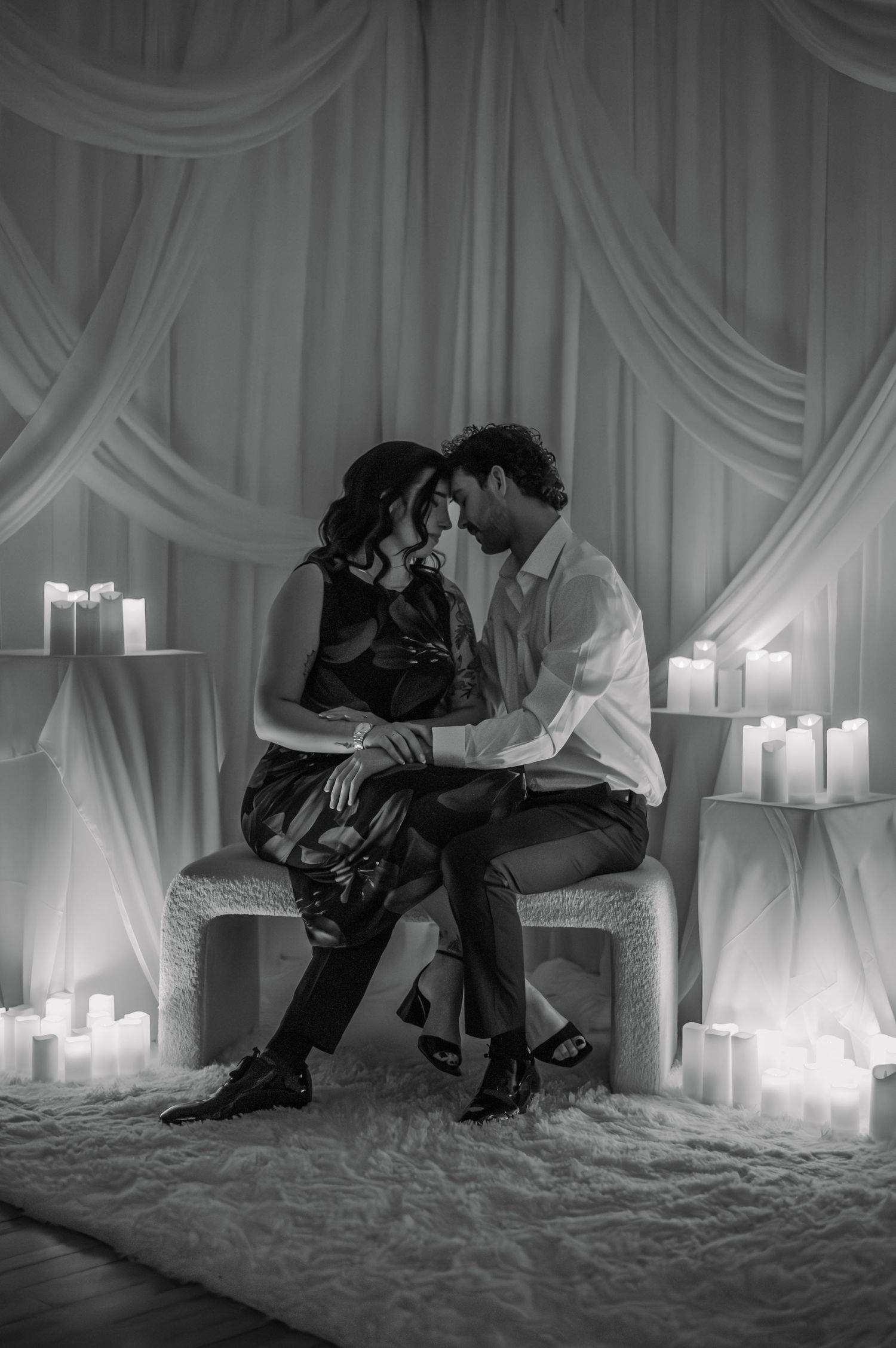 Black and white romantic scene with a couple embracing on a white set with glowing candles in the background.