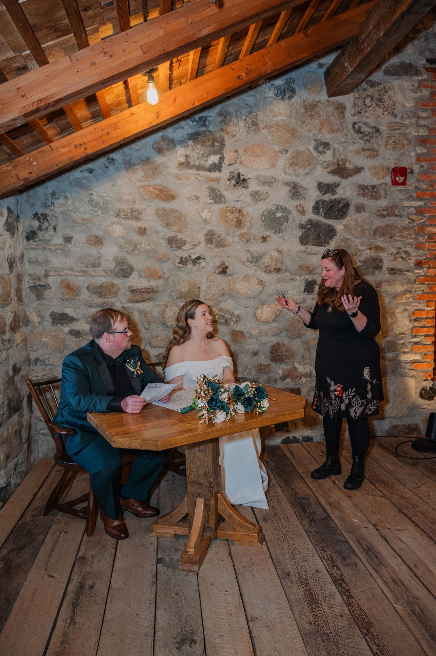 Bride and groom sign wedding documents at wooden table in rustic stone venue with officiant standing nearby.