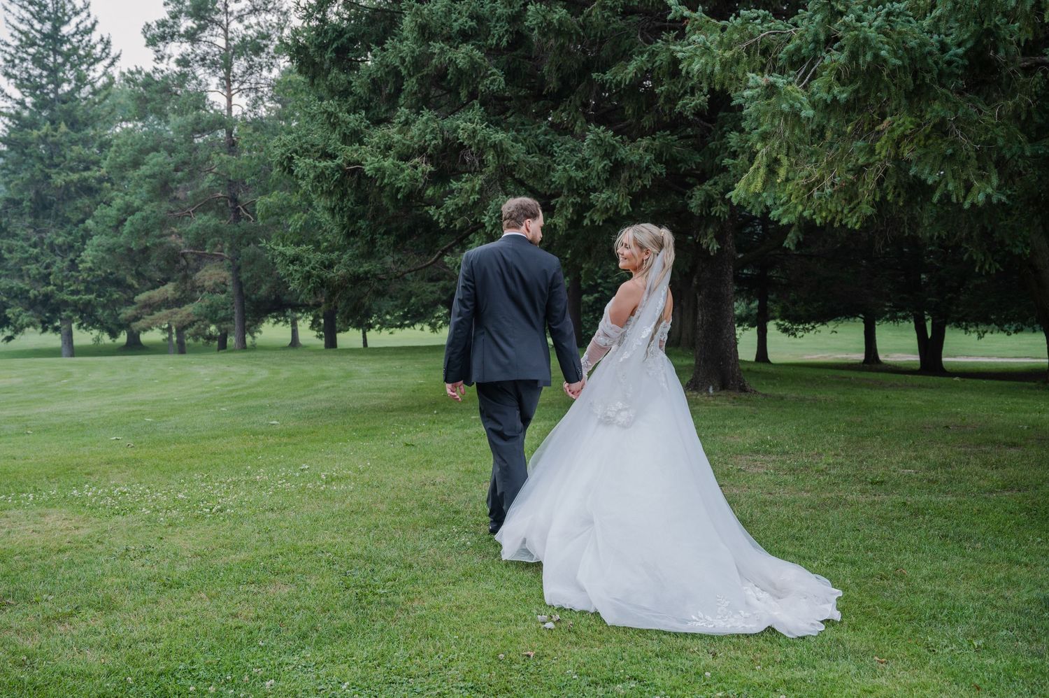 Bride and groom walking hand in hand across lush green lawn surrounded by tall evergreen trees.