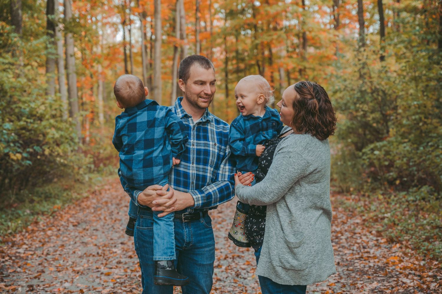 Family shares playful moment on leaf-covered path in autumn forest setting.