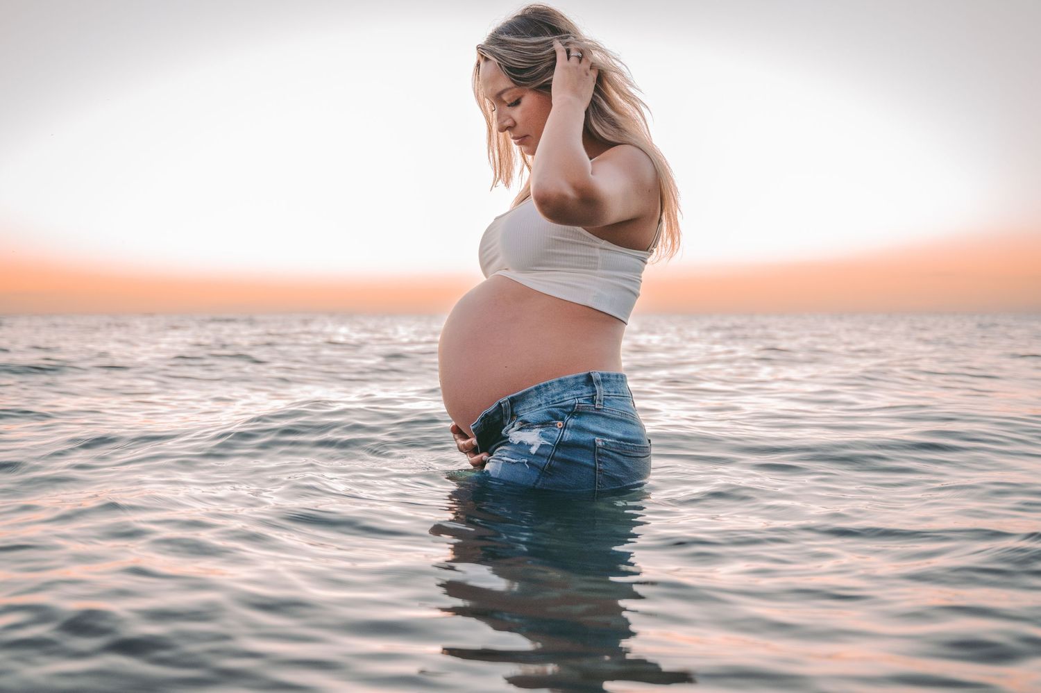 Silhouette of pregnant woman standing in ocean water during sunset maternity shoot.
