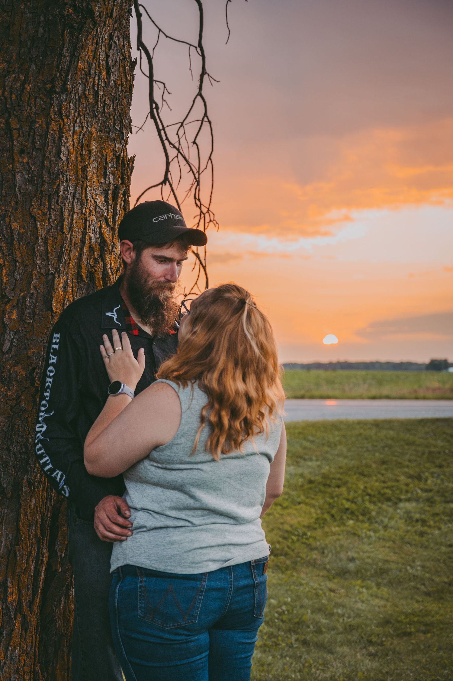 A romantic sunset moment between a couple leaning against a large tree trunk.
