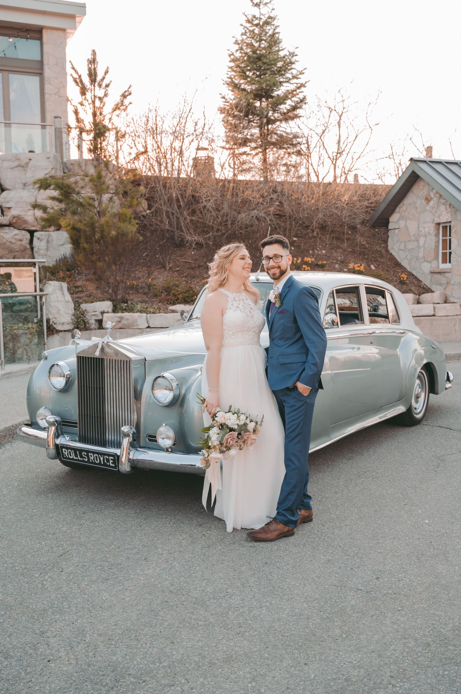 Newlyweds stand beside classic Rolls Royce in elegant wedding attire.