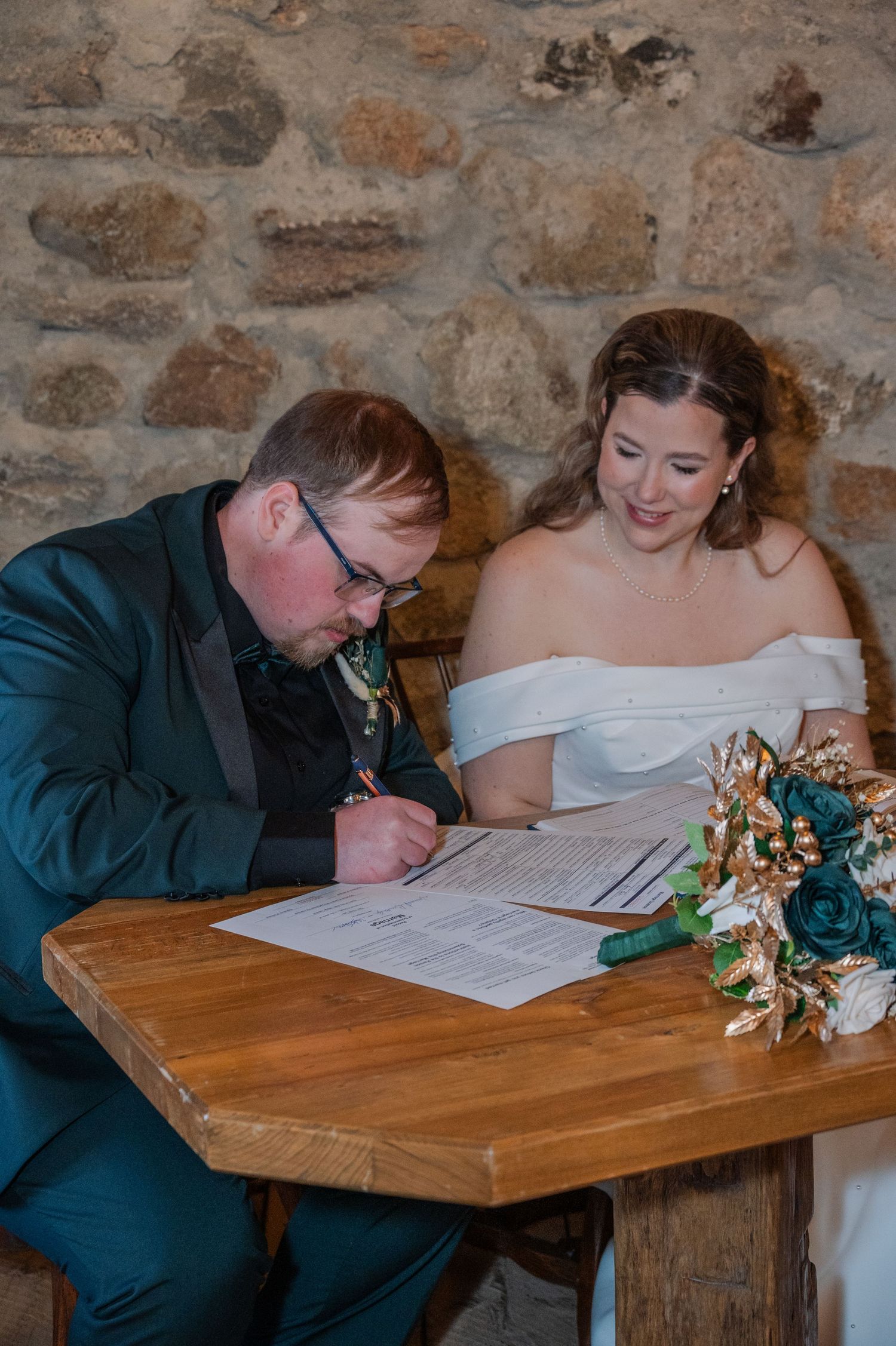 Bride and groom signing wedding marriage certificate at wooden table against rustic stone wall backdrop.