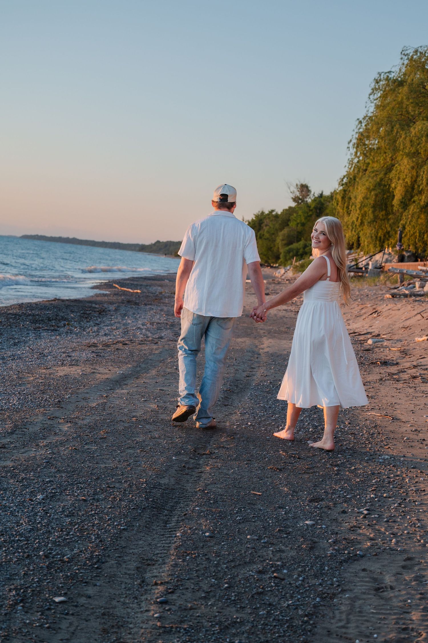 Couple holding hands walking along a rocky beach at sunset, woman in white dress, man in white shirt and jeans.