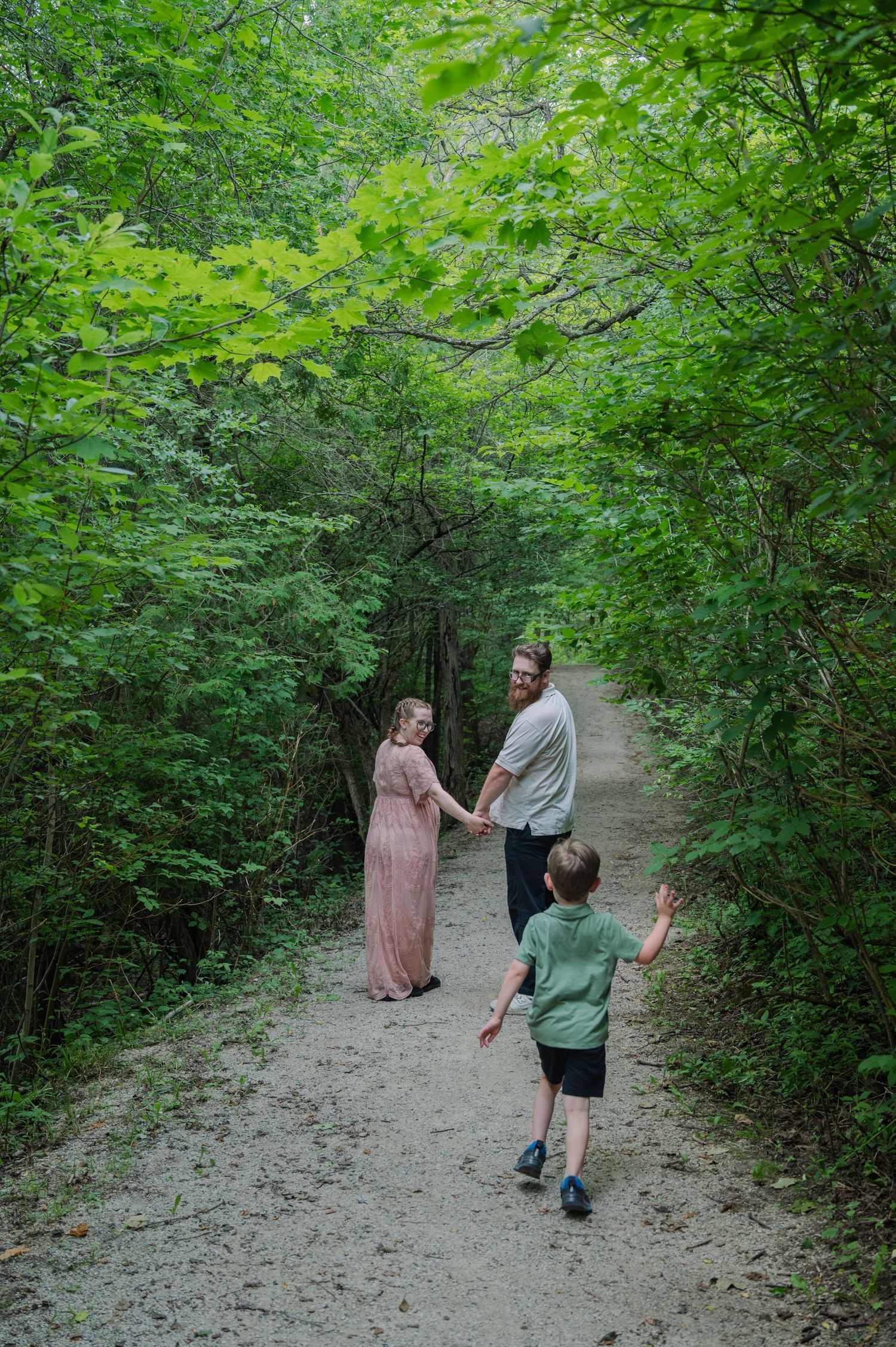 A family walks a lush forest path, parents holding hands as their young son runs toward them.