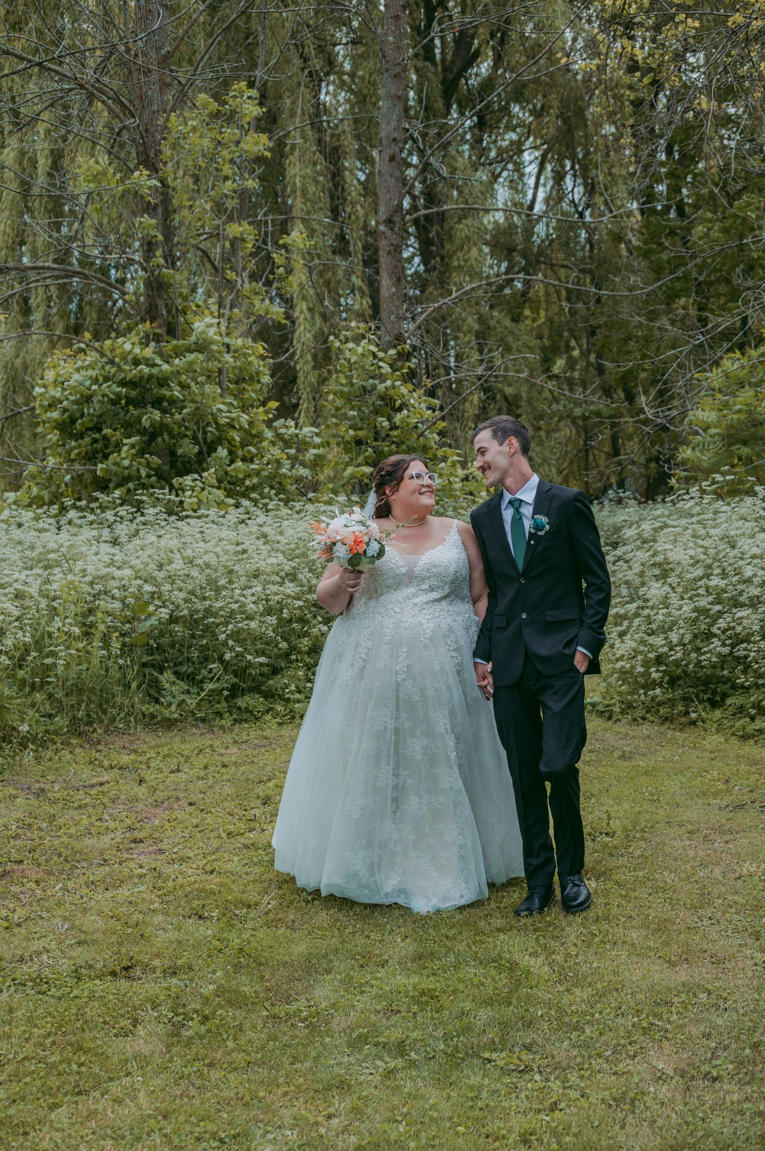 Bride and groom holding hands walking on grass with lush green trees in background at outdoor wedding.
