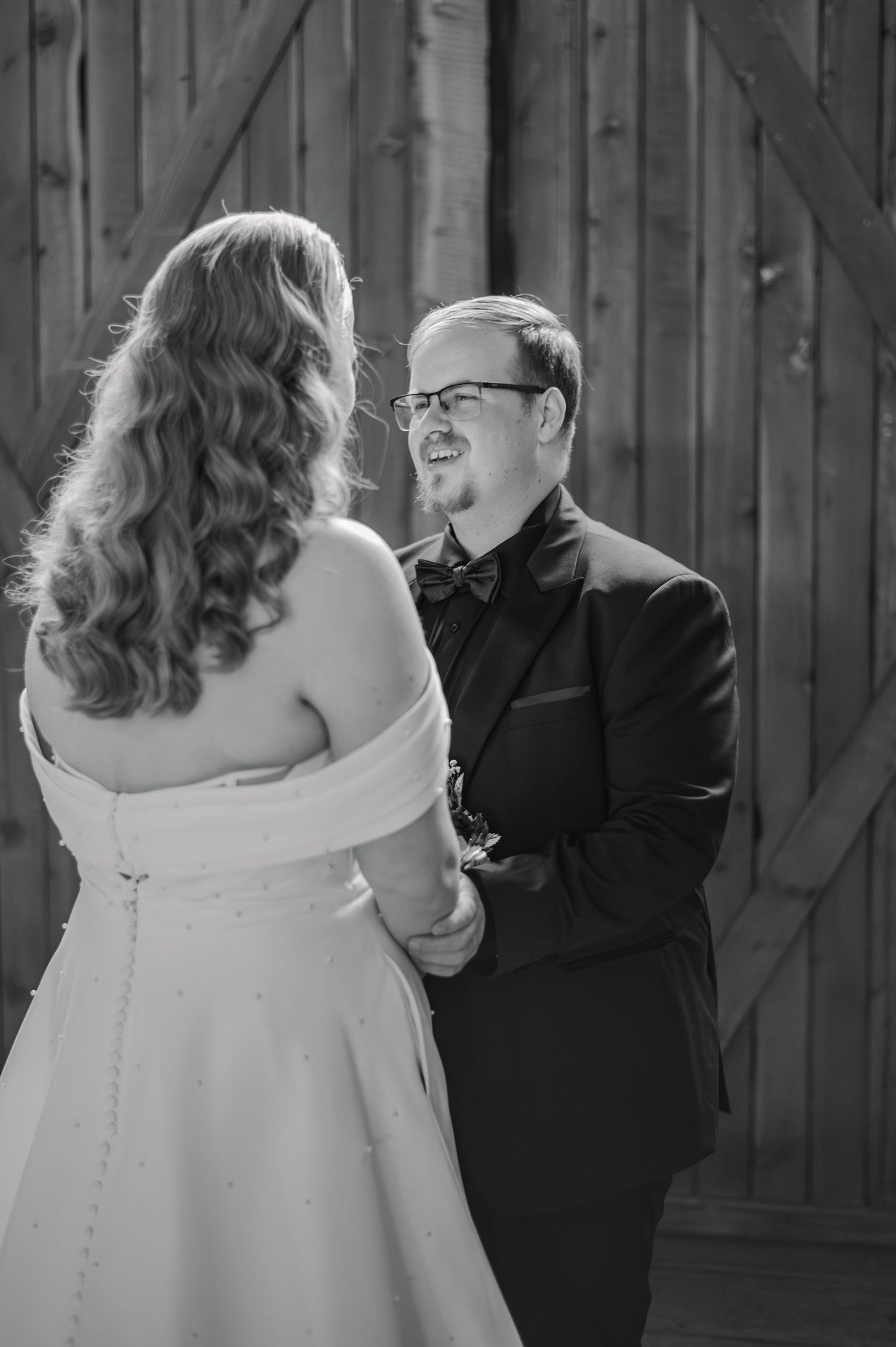 Black and white photo of a bride and groom holding hands, smiling at each other in front of a rustic wooden barn.