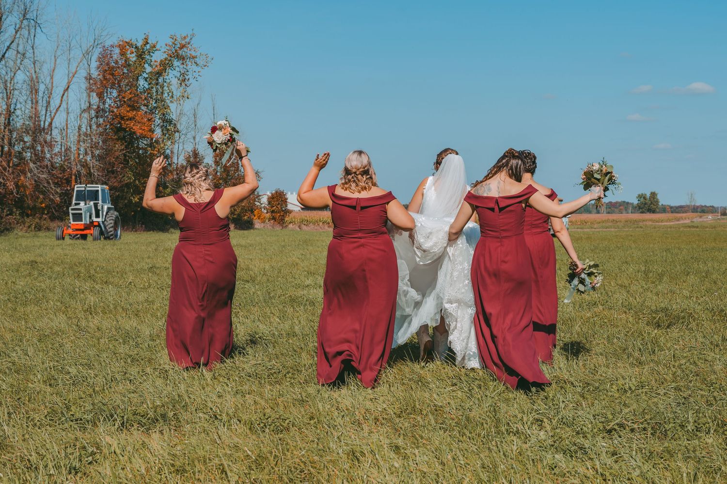 Bridesmaids in burgundy dresses dance and celebrate with bride in white gown against blue sky and autumn field.