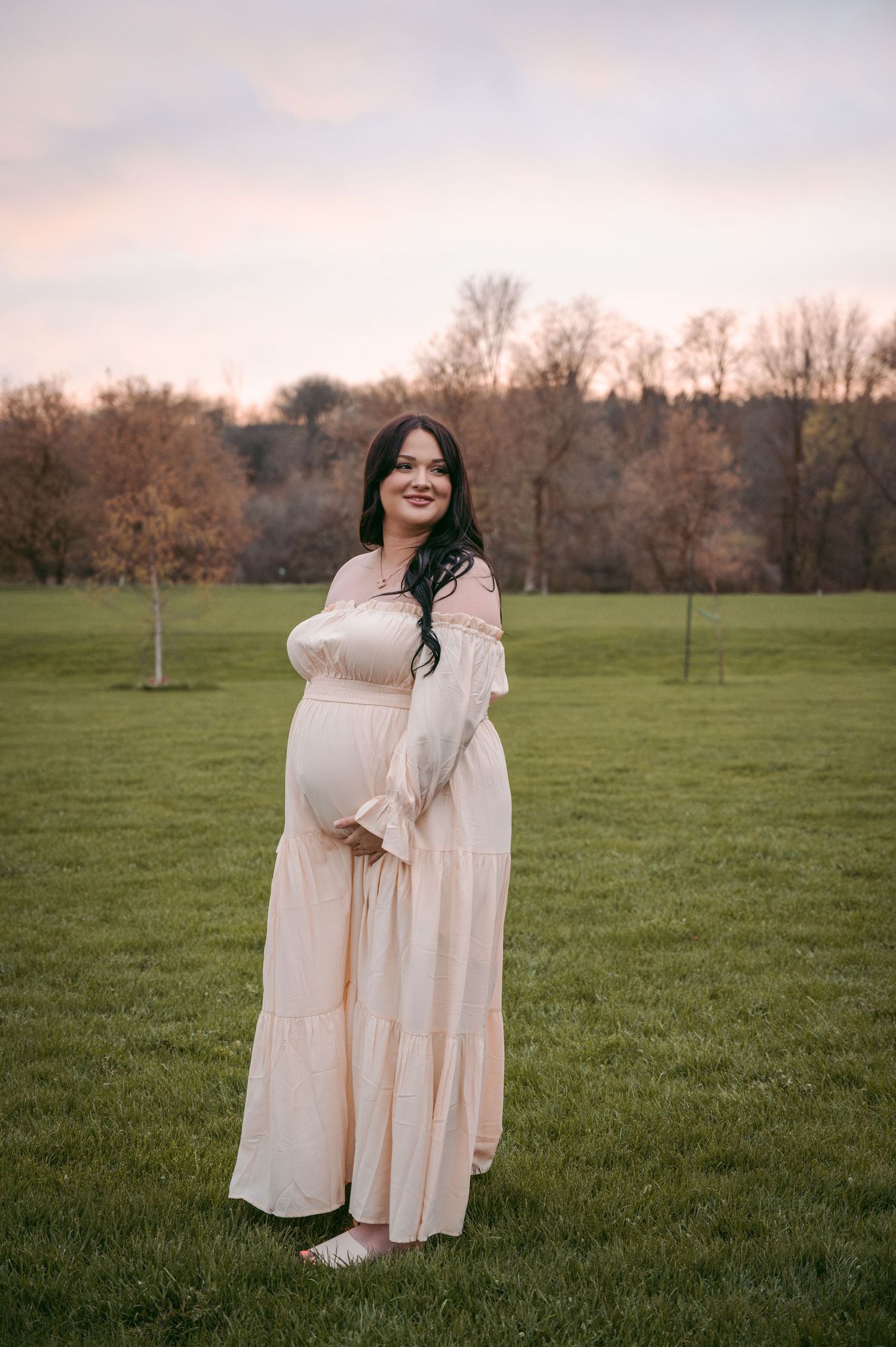 Pregnant woman in flowing cream dress poses in grassy field during golden hour.
