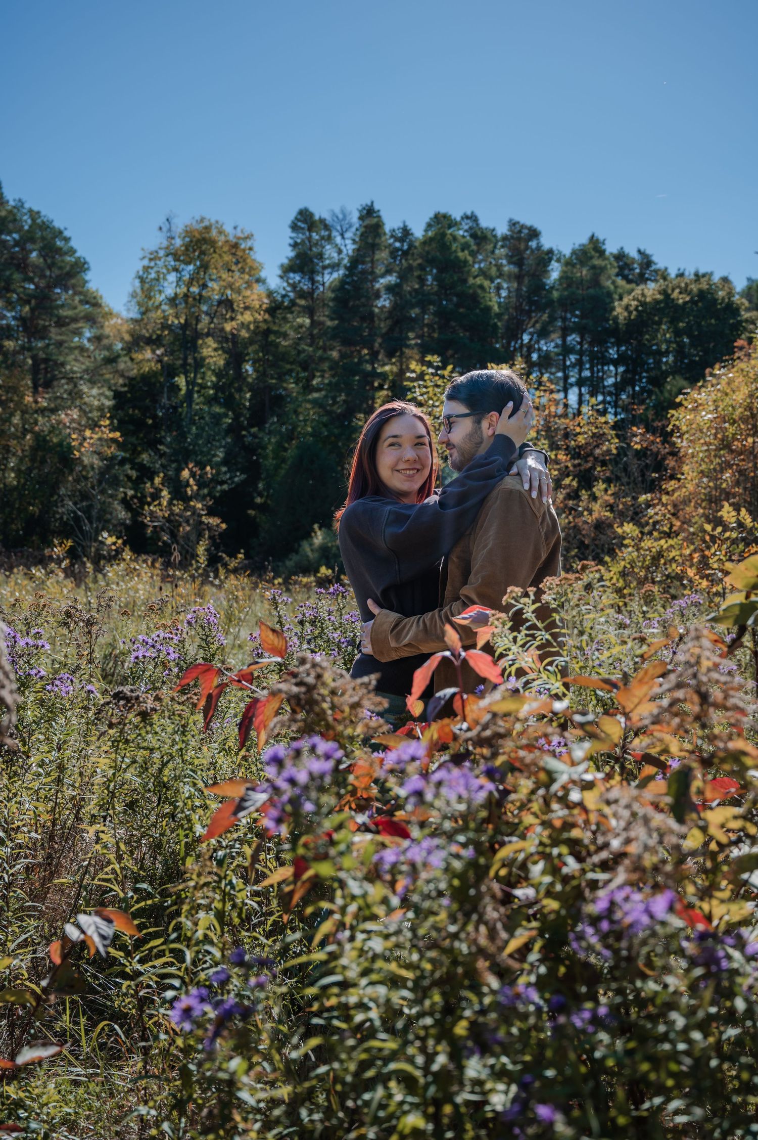 A couple embraces in a vibrant wildflower field with purple and orange blooms against a backdrop of tall pine trees.