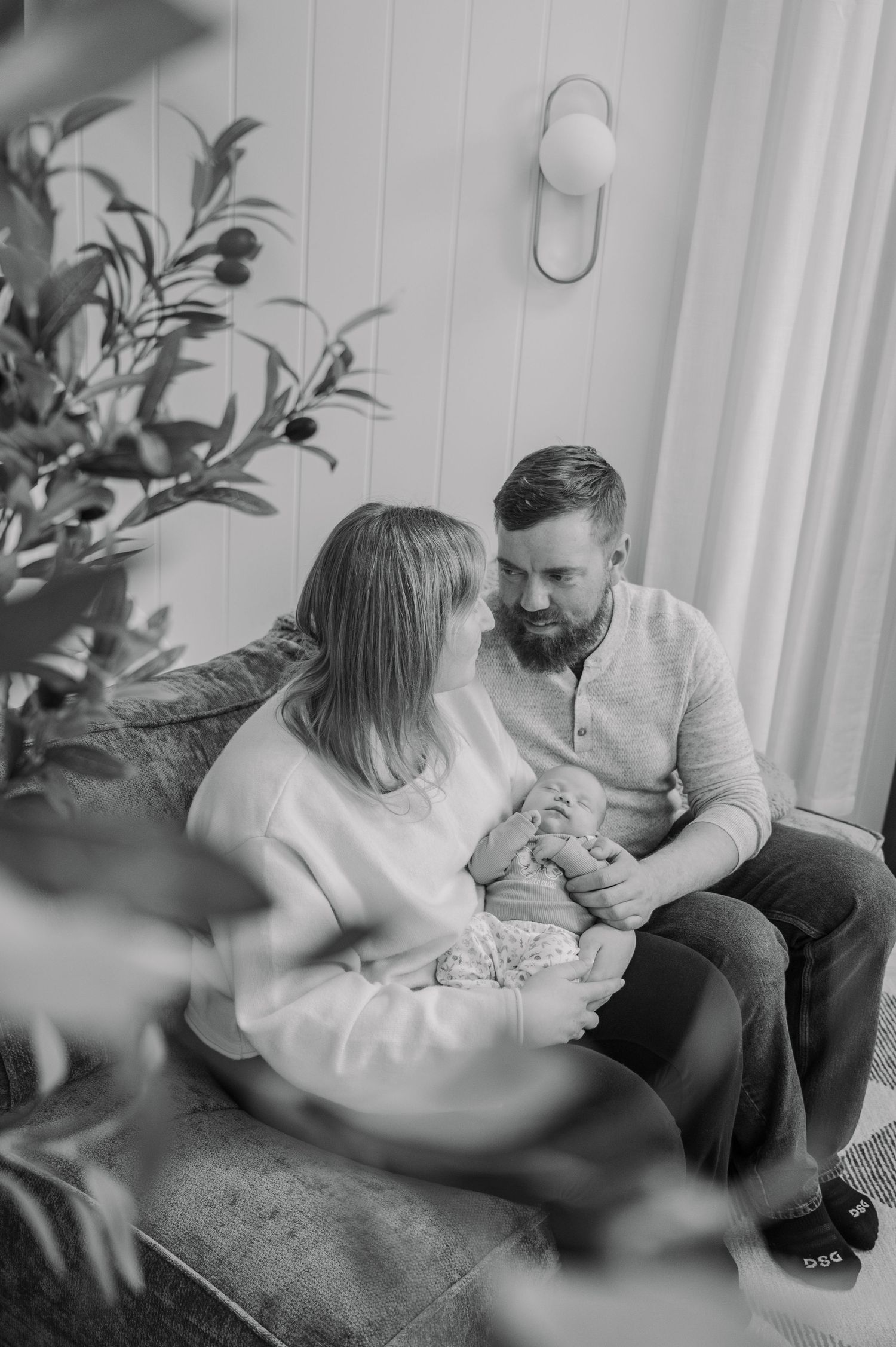 Black and white photo of a couple sitting on a couch, lovingly gazing at their newborn baby.