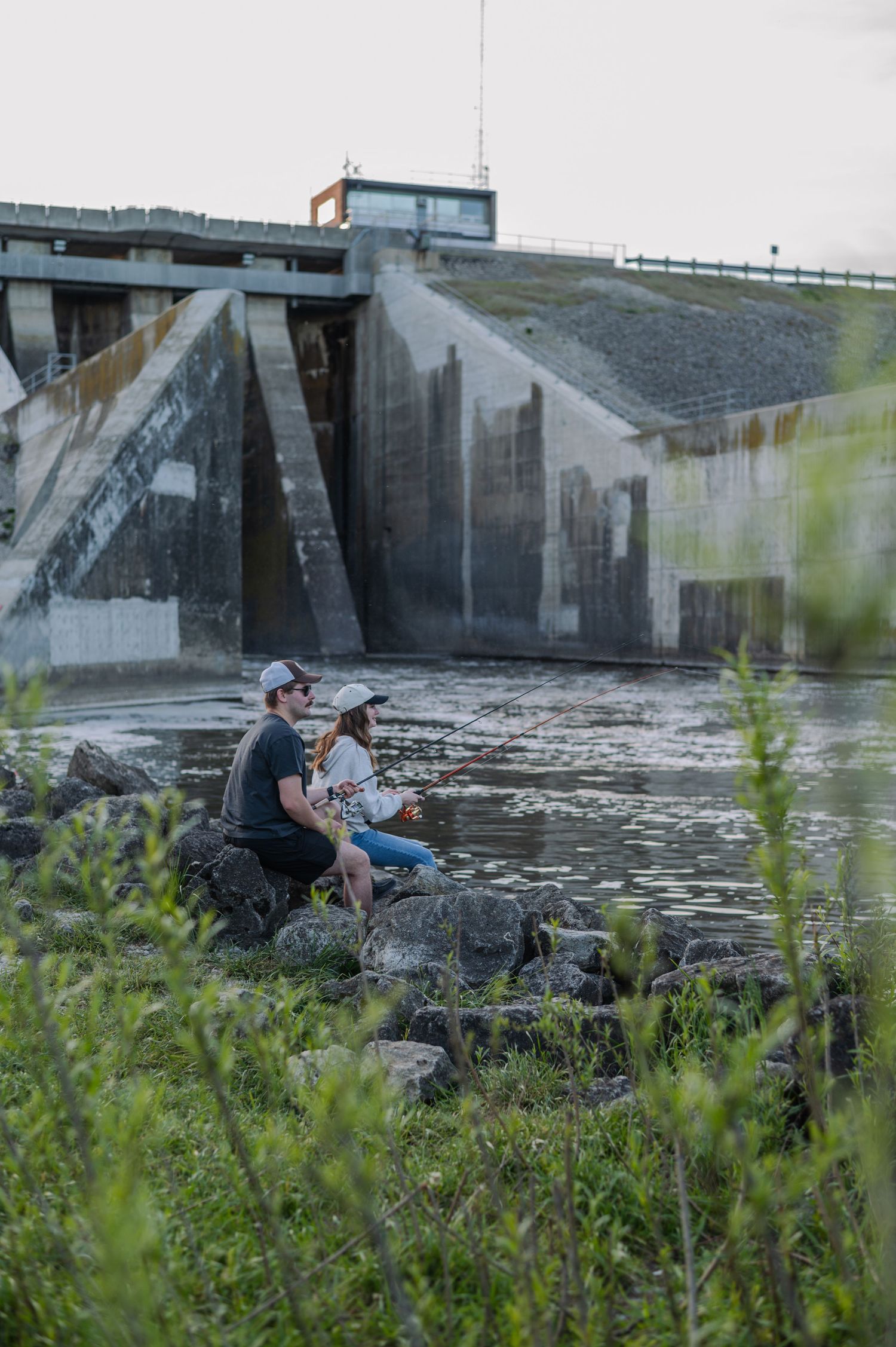 Two people fishing on rocks near a dam spillway, surrounded by lush greenery and flowing water.