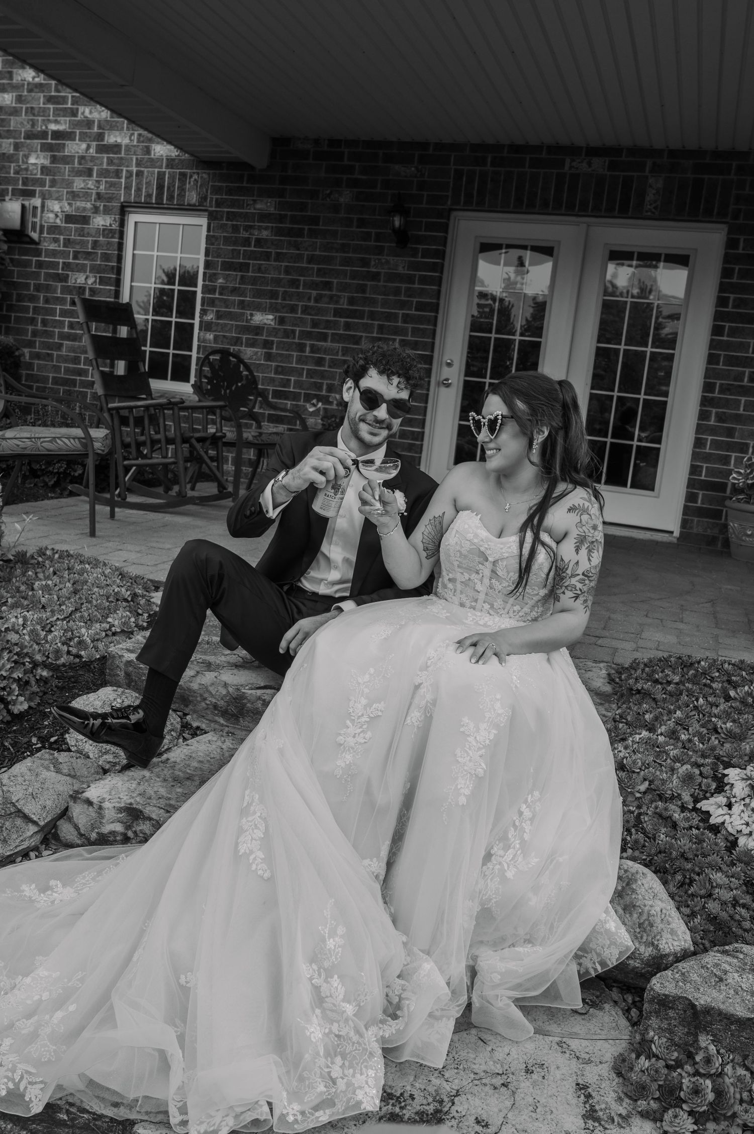 Black and white photo of a bride and groom in sunglasses sitting on stone steps outside a brick home.