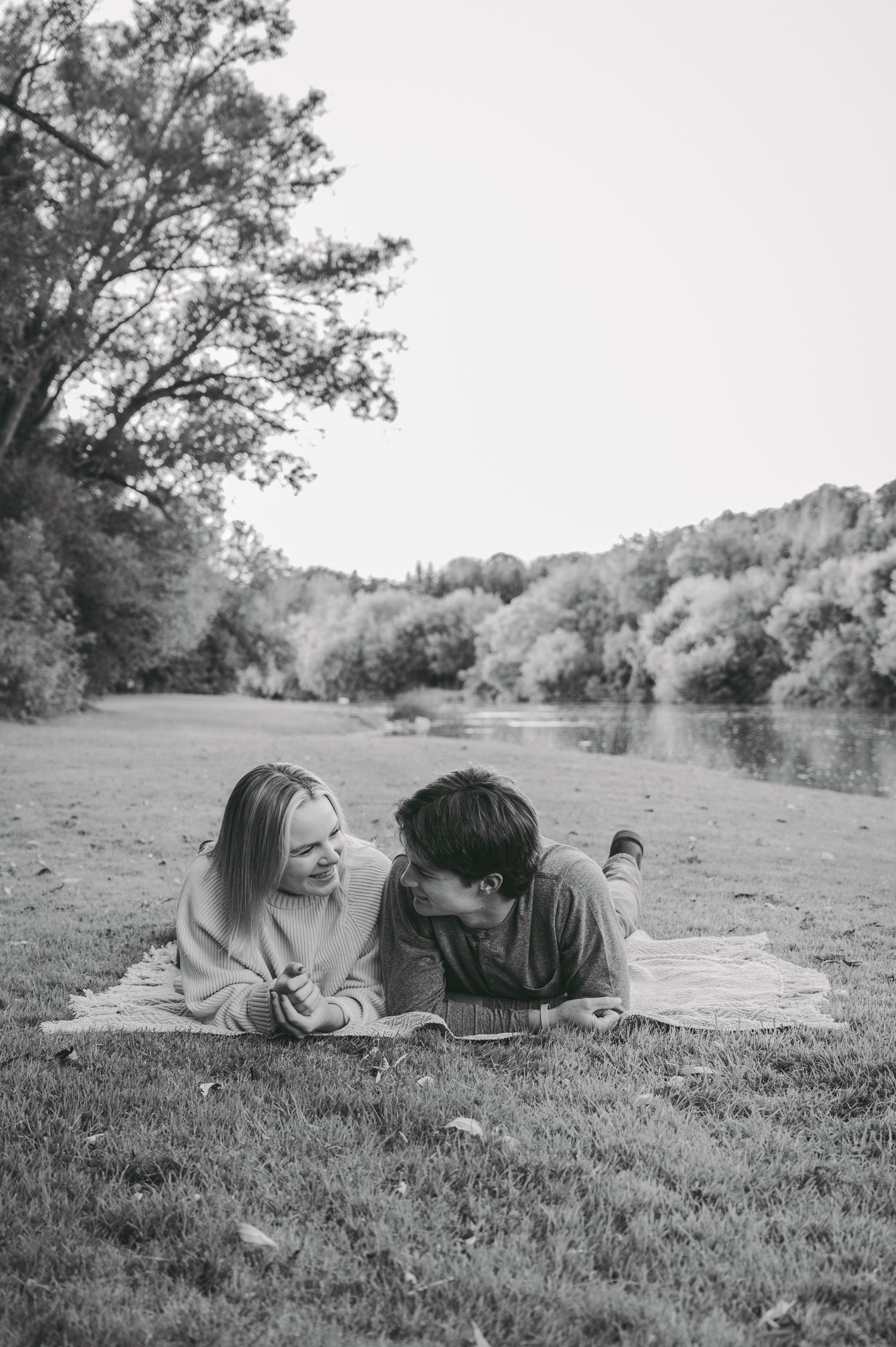 A couple relaxes on a blanket by a lake in a black and white photograph.