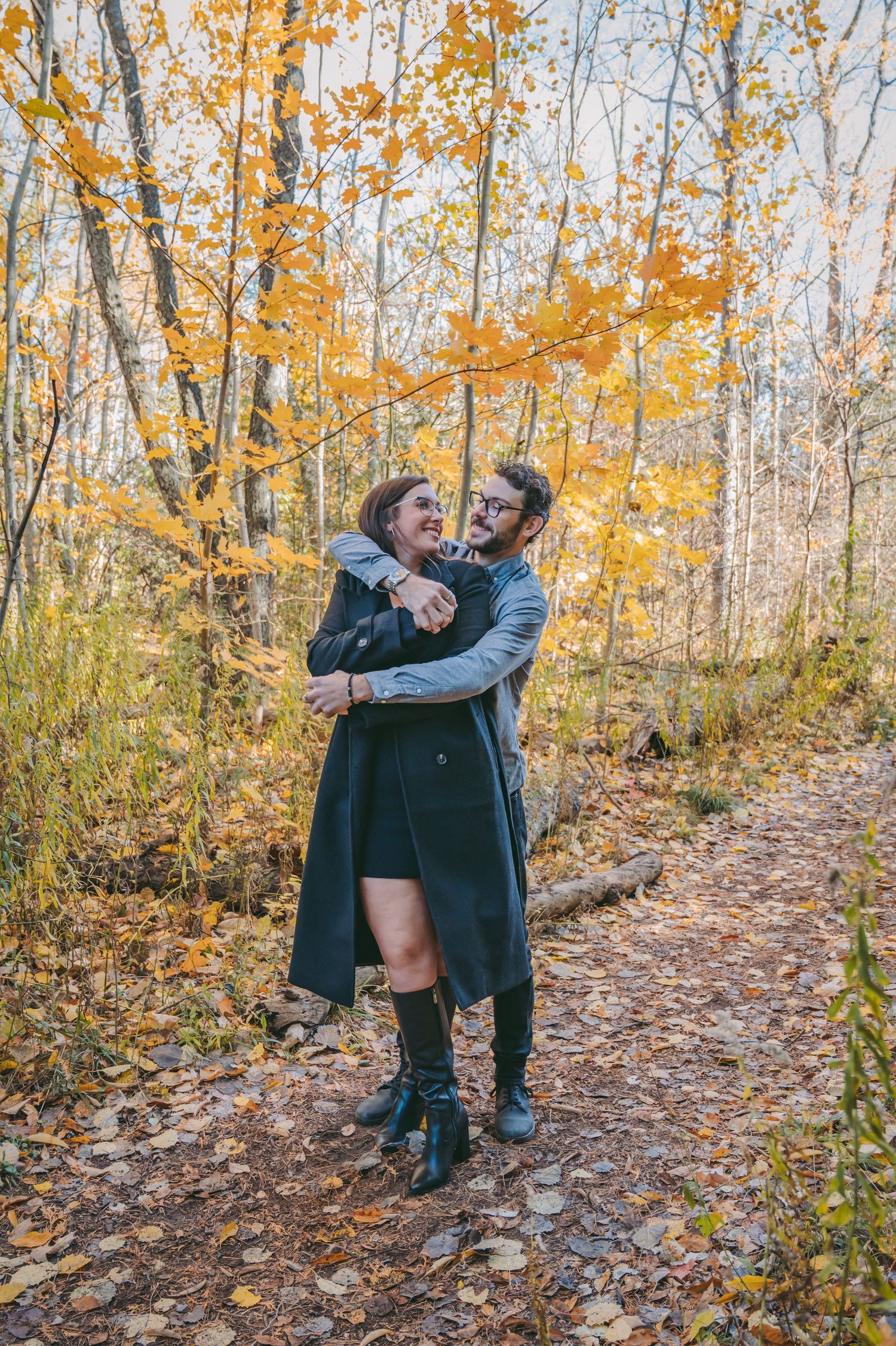 A couple embraces on a forest trail surrounded by golden autumn foliage and fallen leaves.