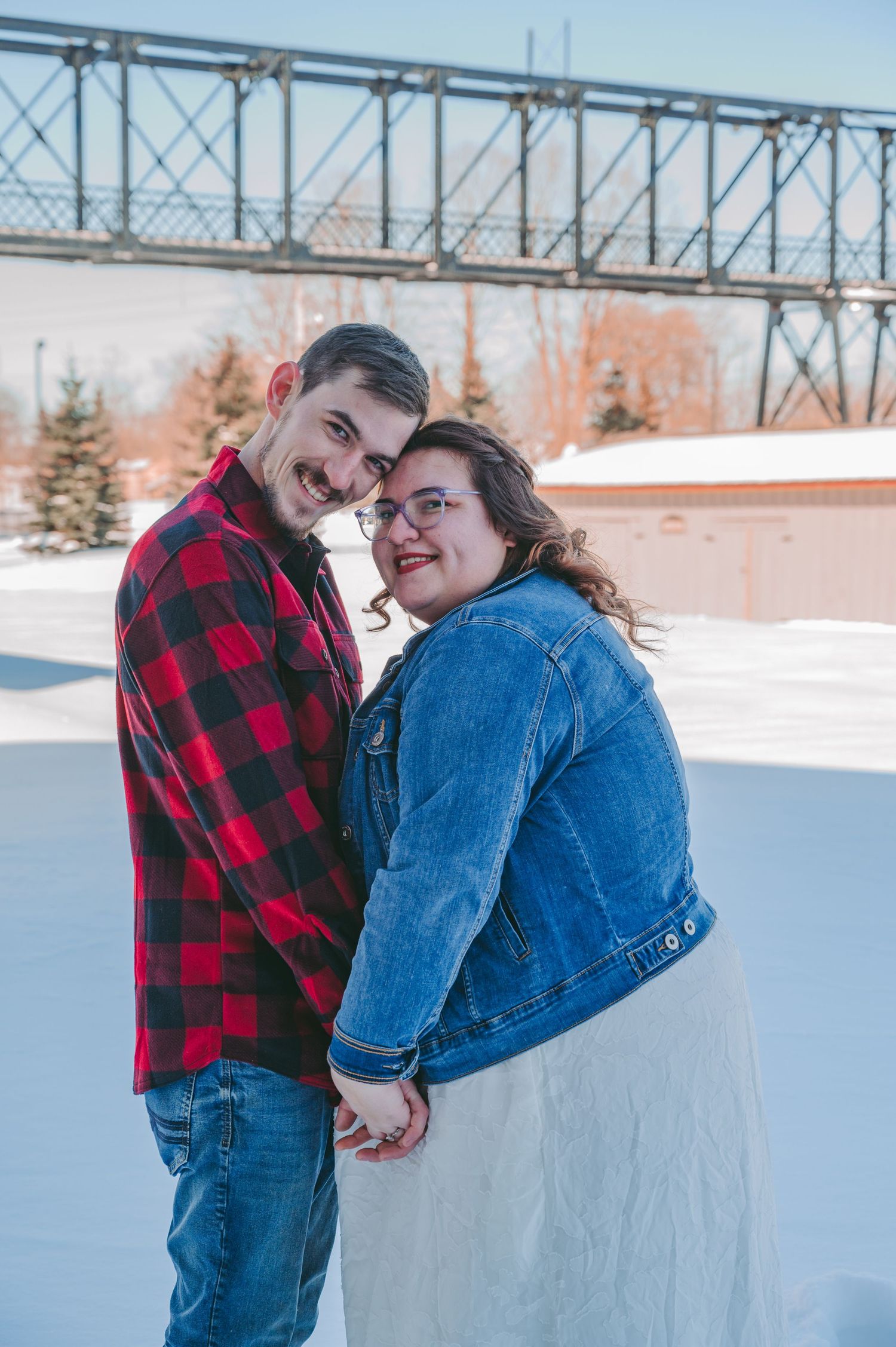 Couple poses playfully in winter setting with metal bridge structure visible.