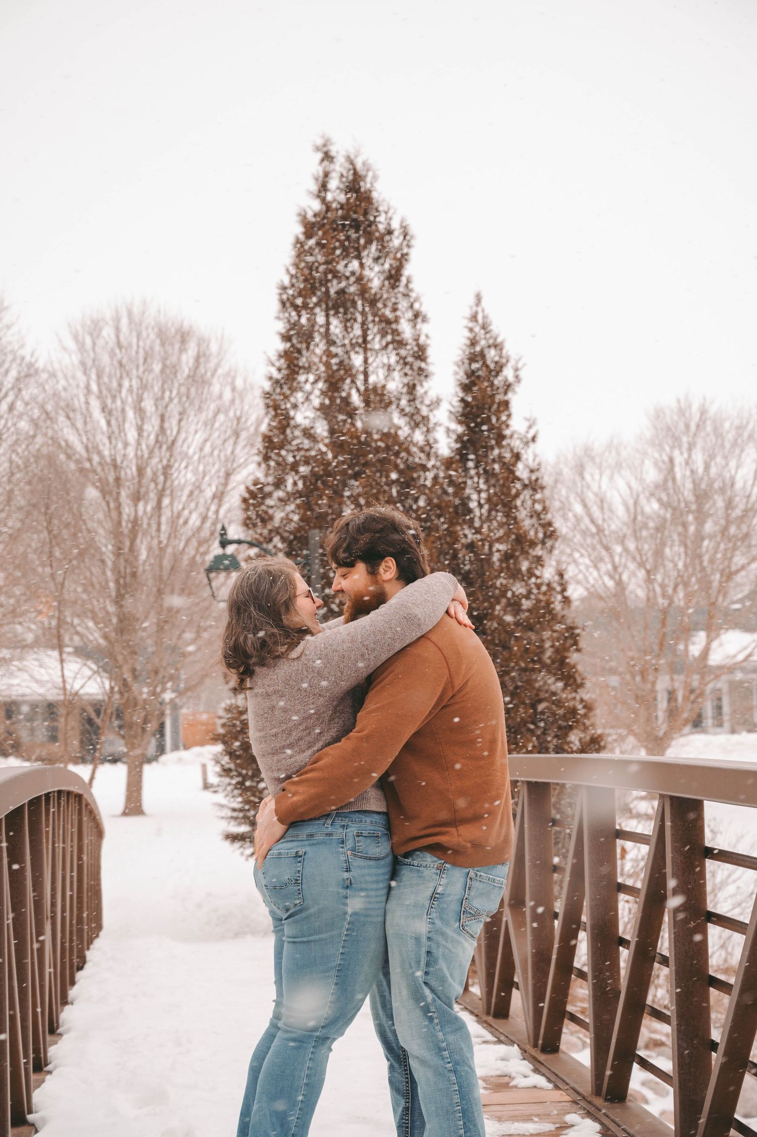 Couple embracing on snowy bridge with evergreen trees in winter landscape.