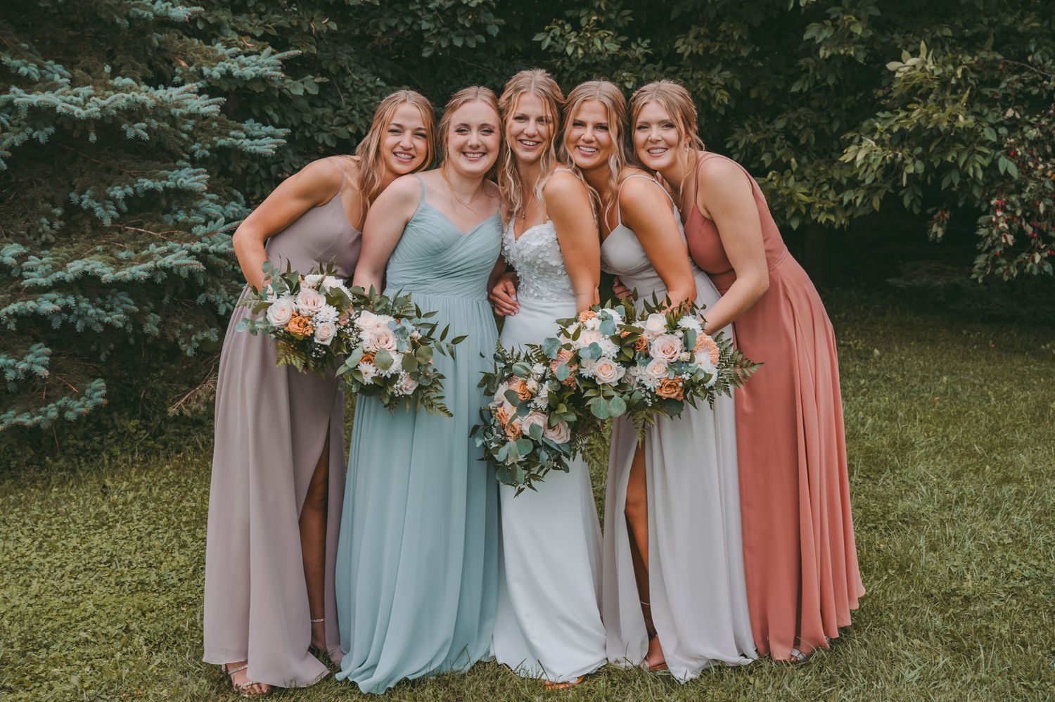 Bridesmaids in pastel dresses holding floral bouquets pose together on lawn.