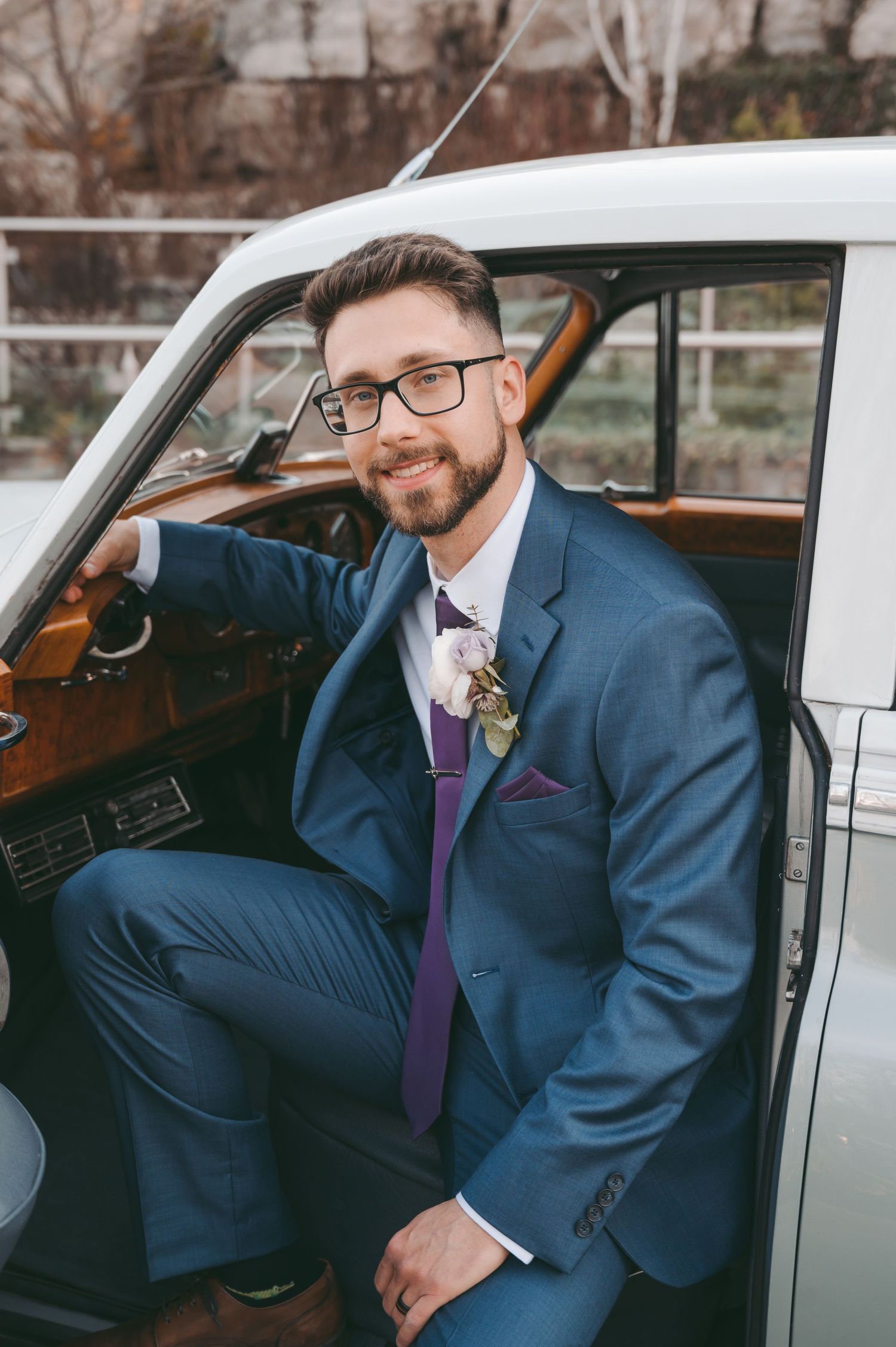 Wedding party member in a blue suit and purple tie seated in classic vehicle.