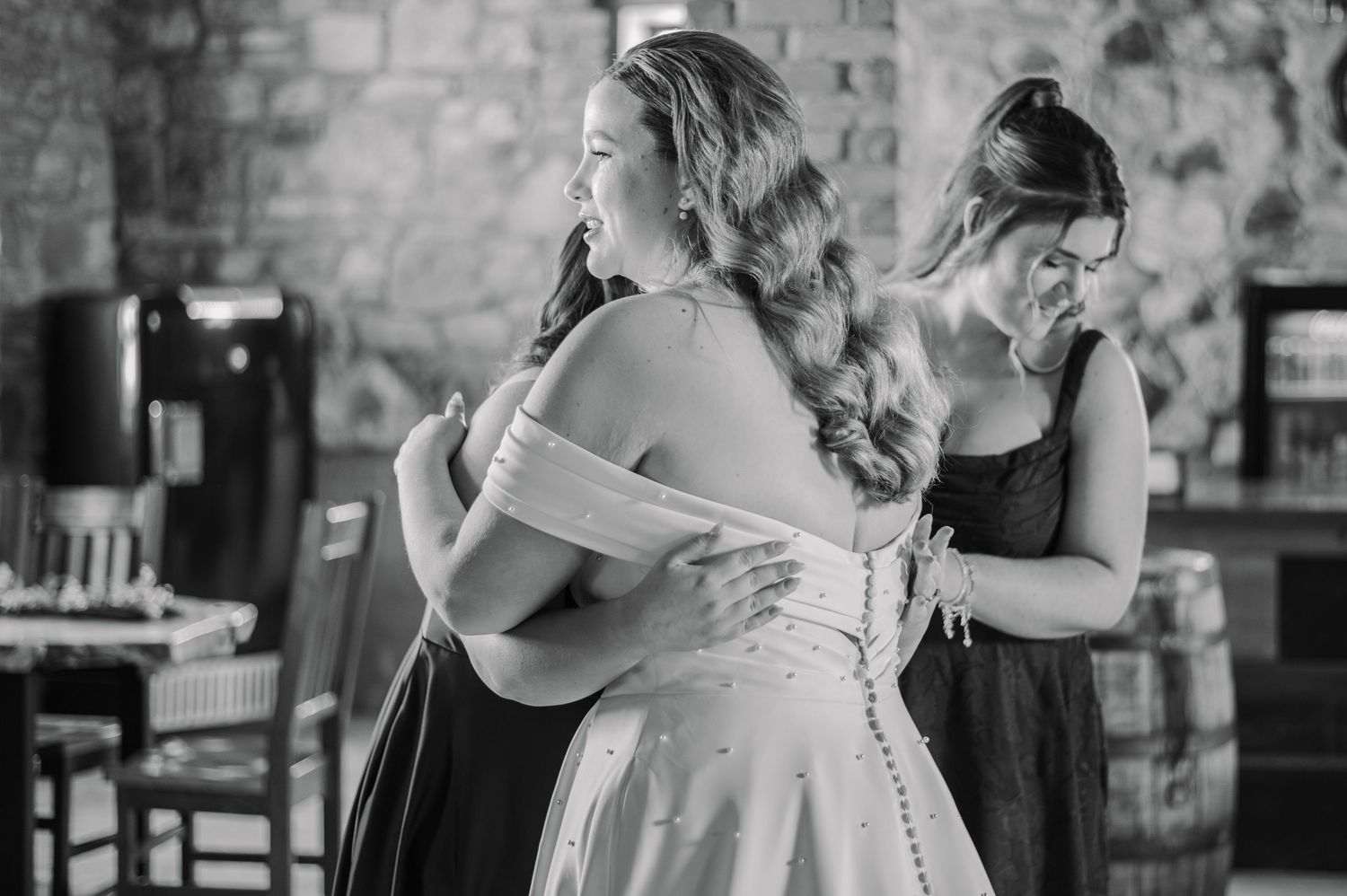 Bride hugging bridesmaid while another helps button her wedding dress in a rustic venue, black and white photo.