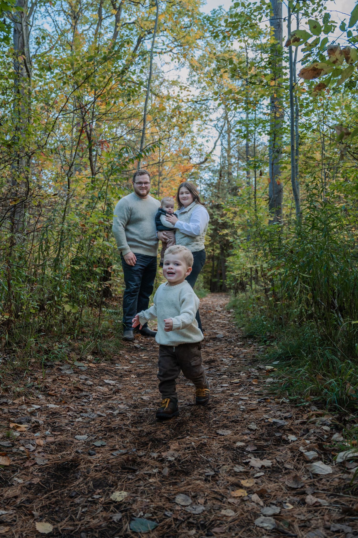 Family enjoying an autumn walk together on a leaf-covered forest trail surrounded by tall trees and greenery.