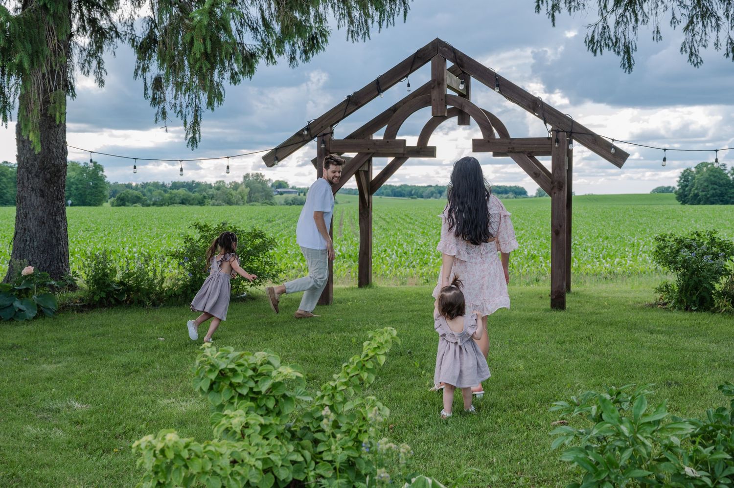 Family playing near a wooden pergola surrounded by lush green fields and trees on a cloudy day.