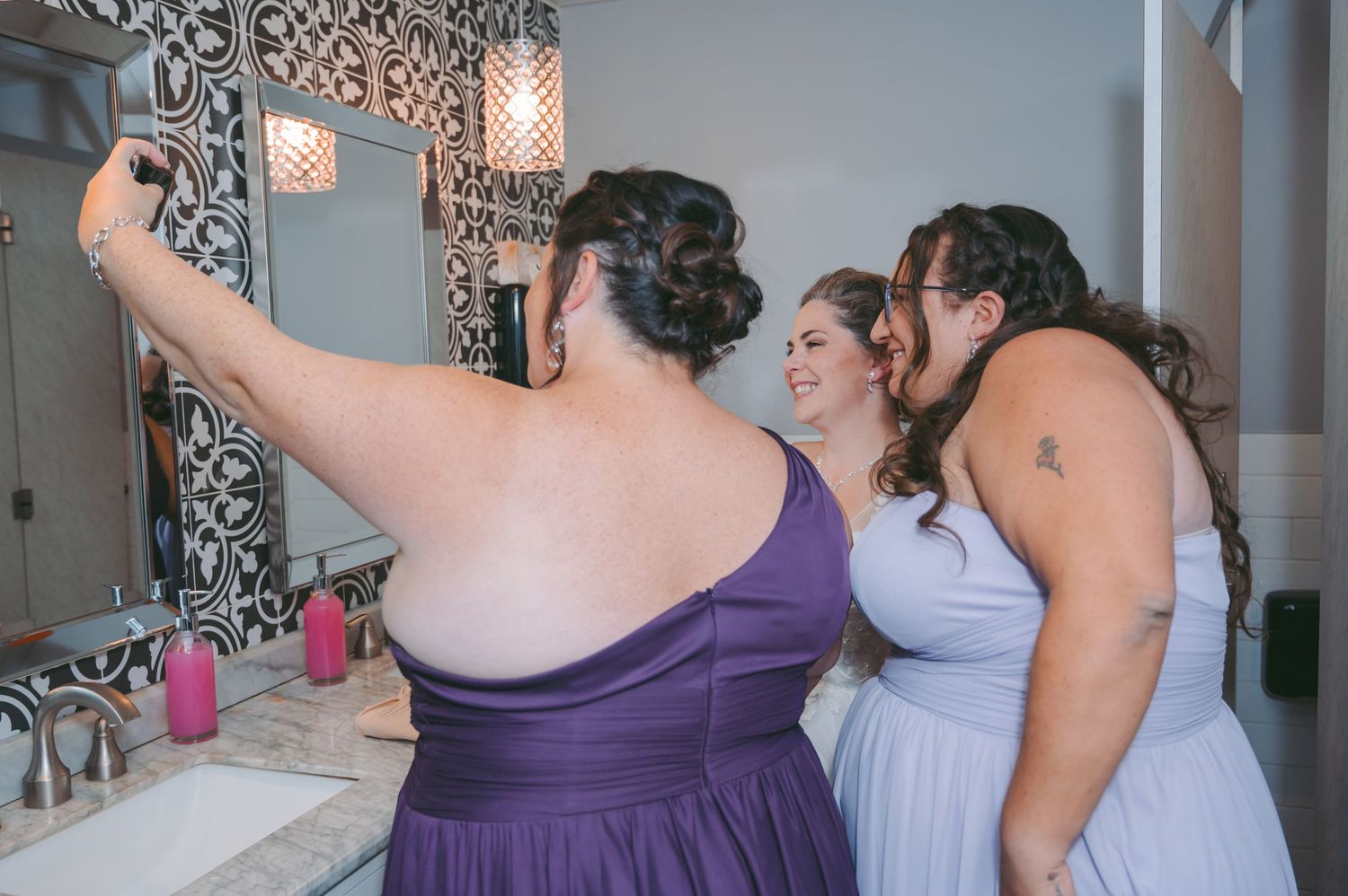 Bridesmaids in purple dresses taking selfies in a bathroom with decorative lighting.