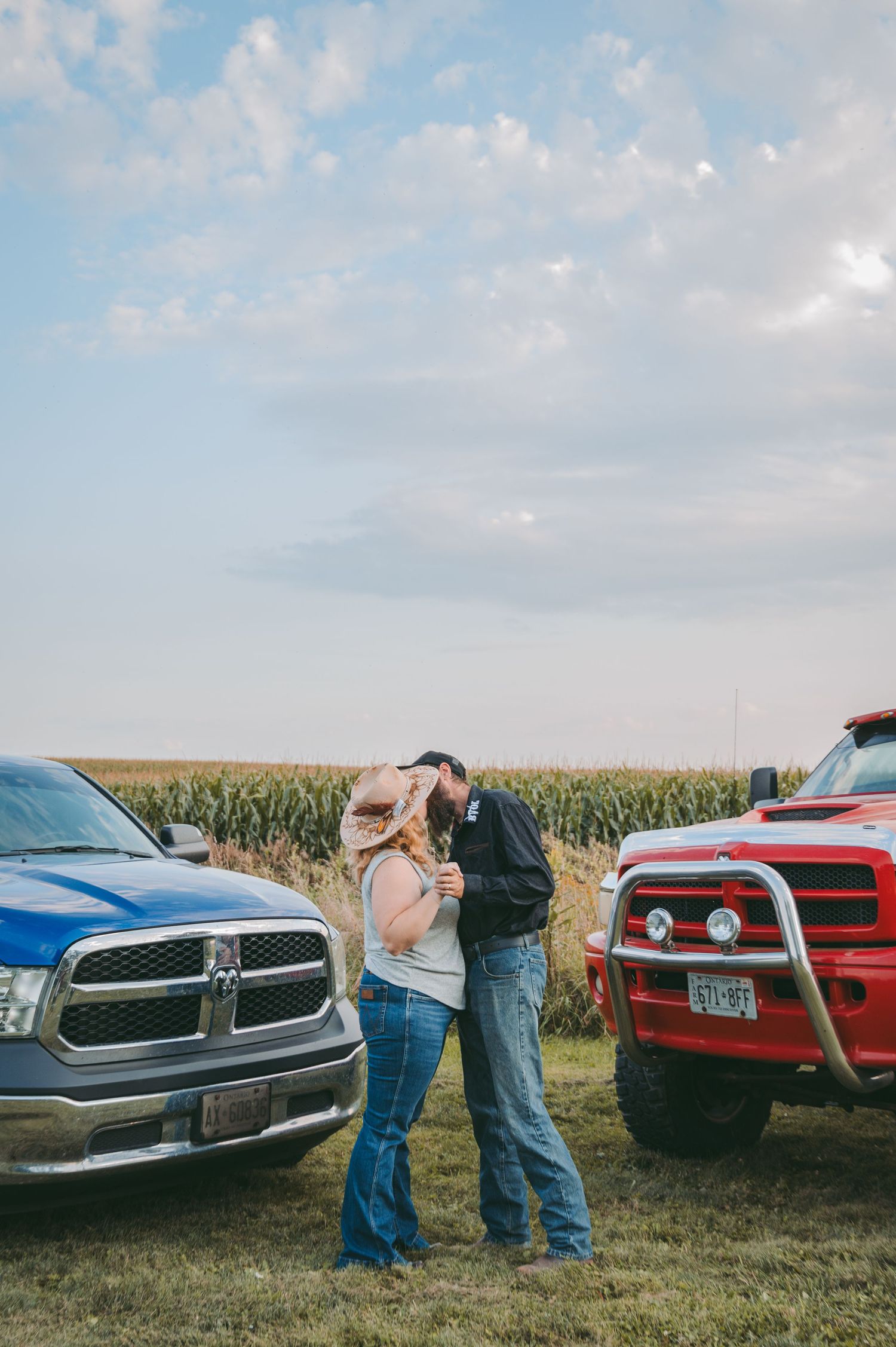 A couple shares an intimate moment between two pickup trucks in a cornfield at sunset.