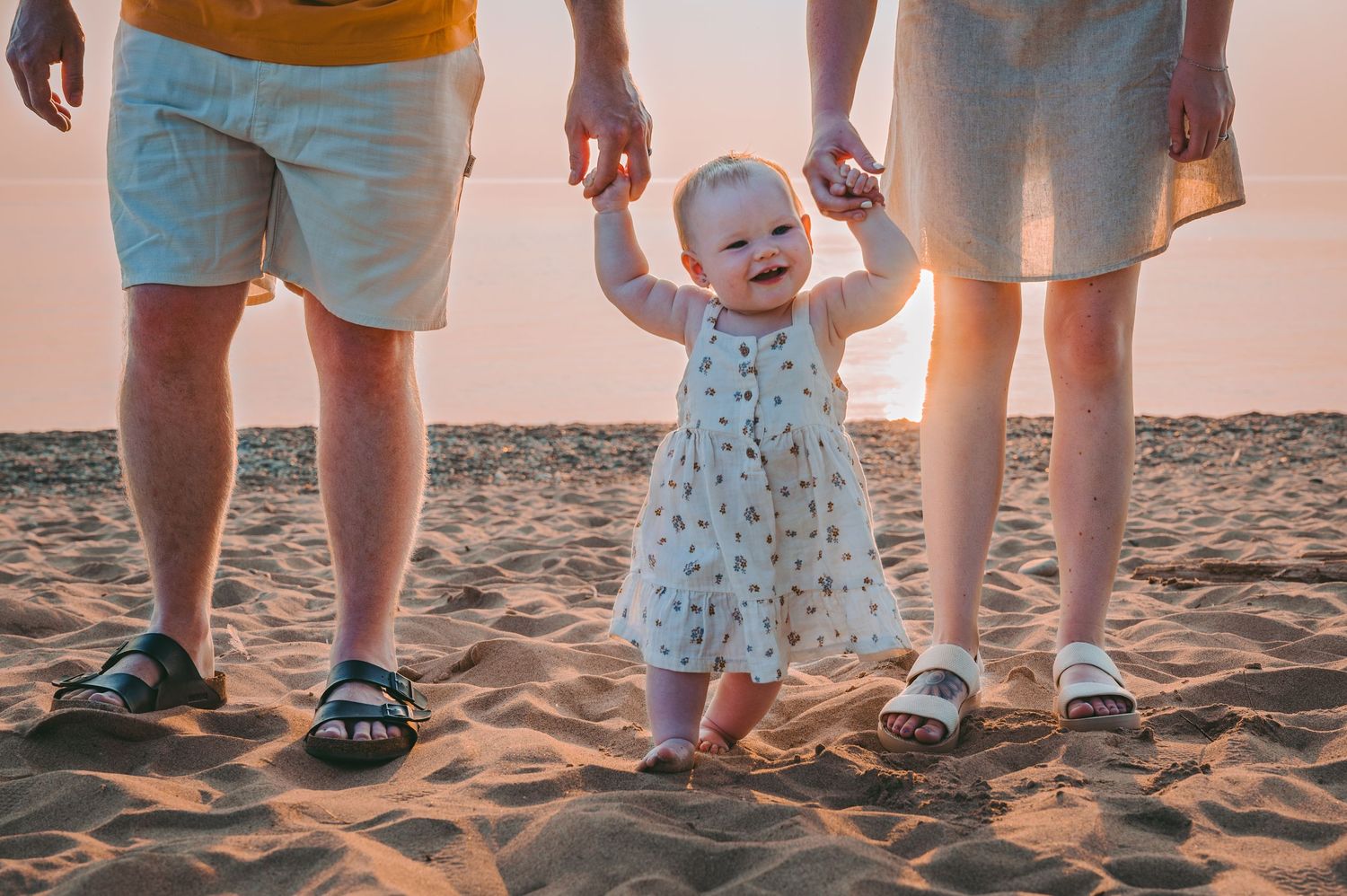 A small child takes first steps on sandy beach at sunset while holding hands with parents.