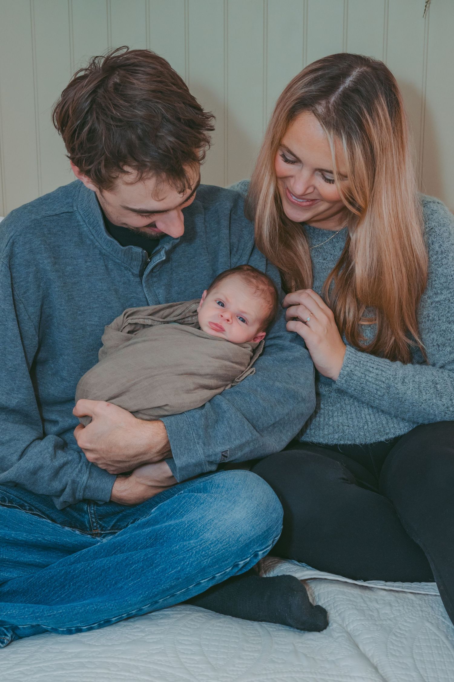 New parents in cozy sweaters cradle their swaddled newborn baby on white bedding.