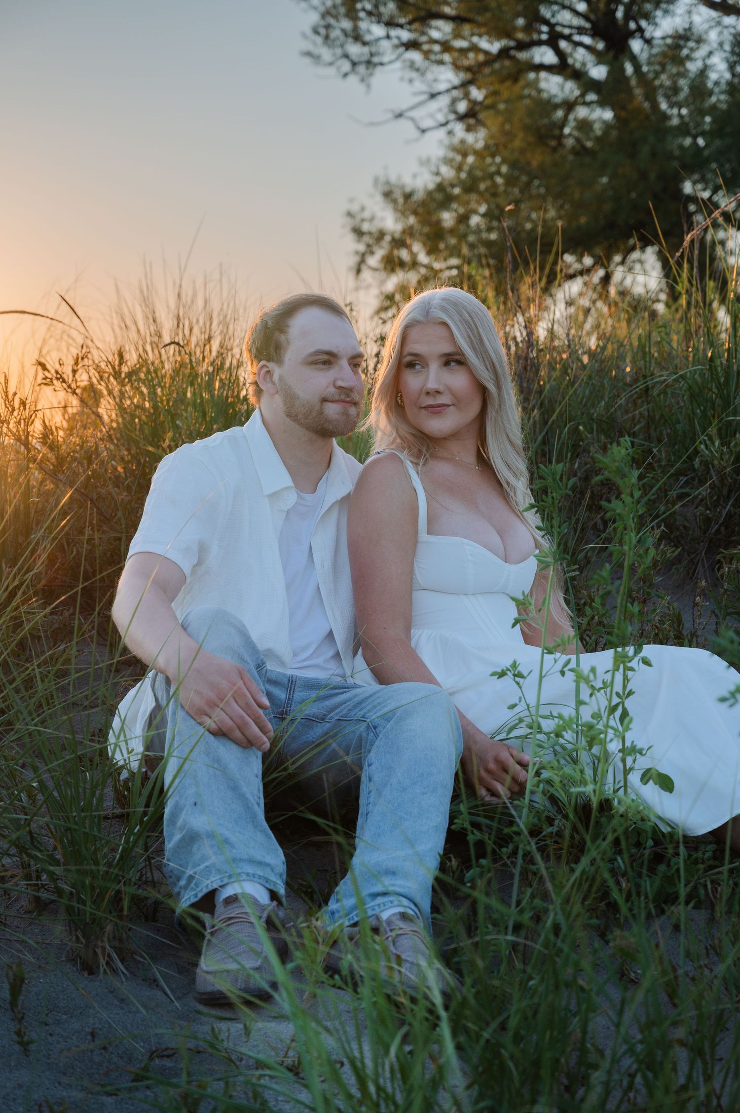 Couple sitting together in golden hour sunset among tall grass and dunes, wearing white outfits, romantic outdoor portrait.